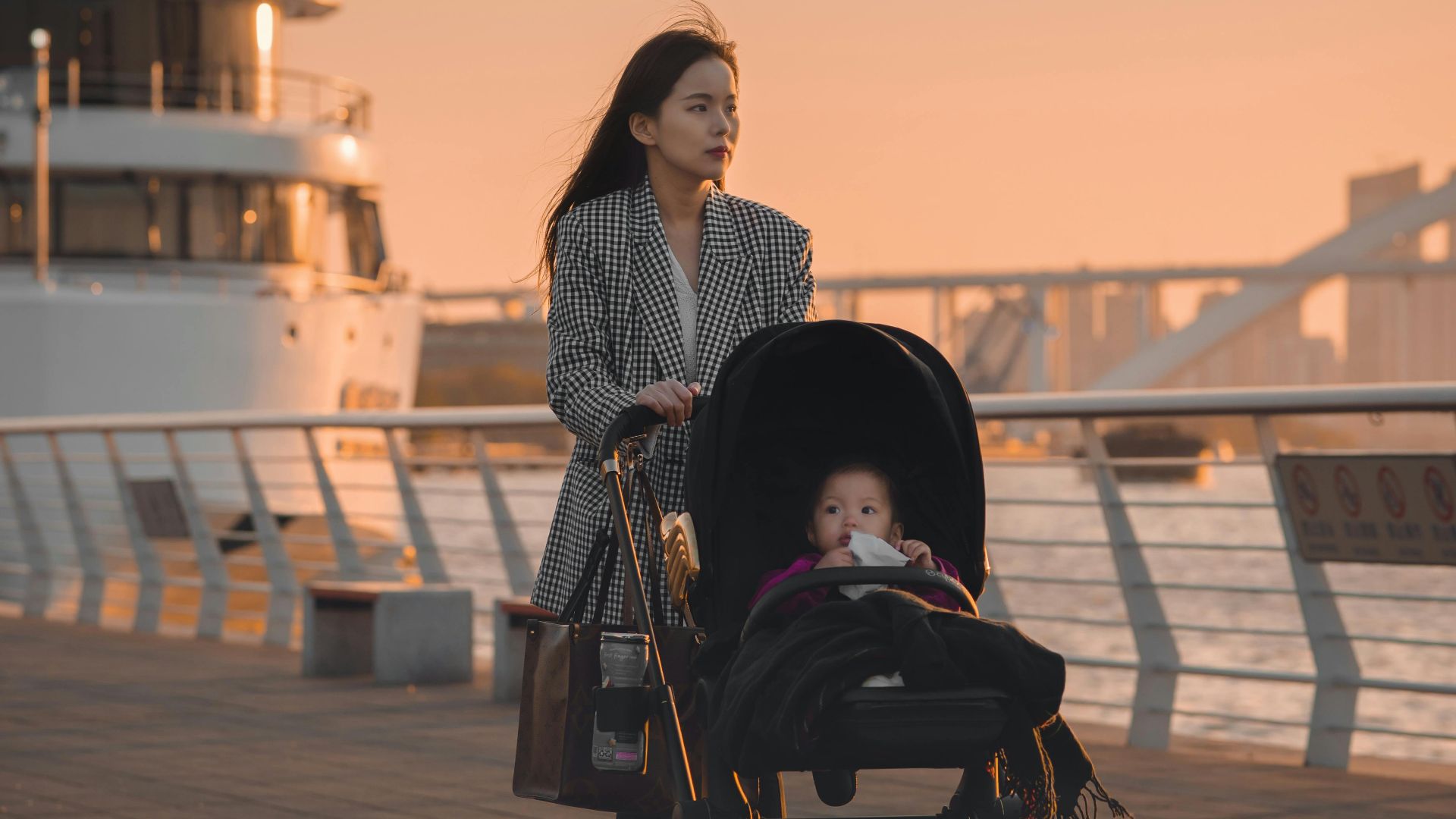 Woman strolling with a baby in a stroller by the pier at sunset, with a ship in the background.