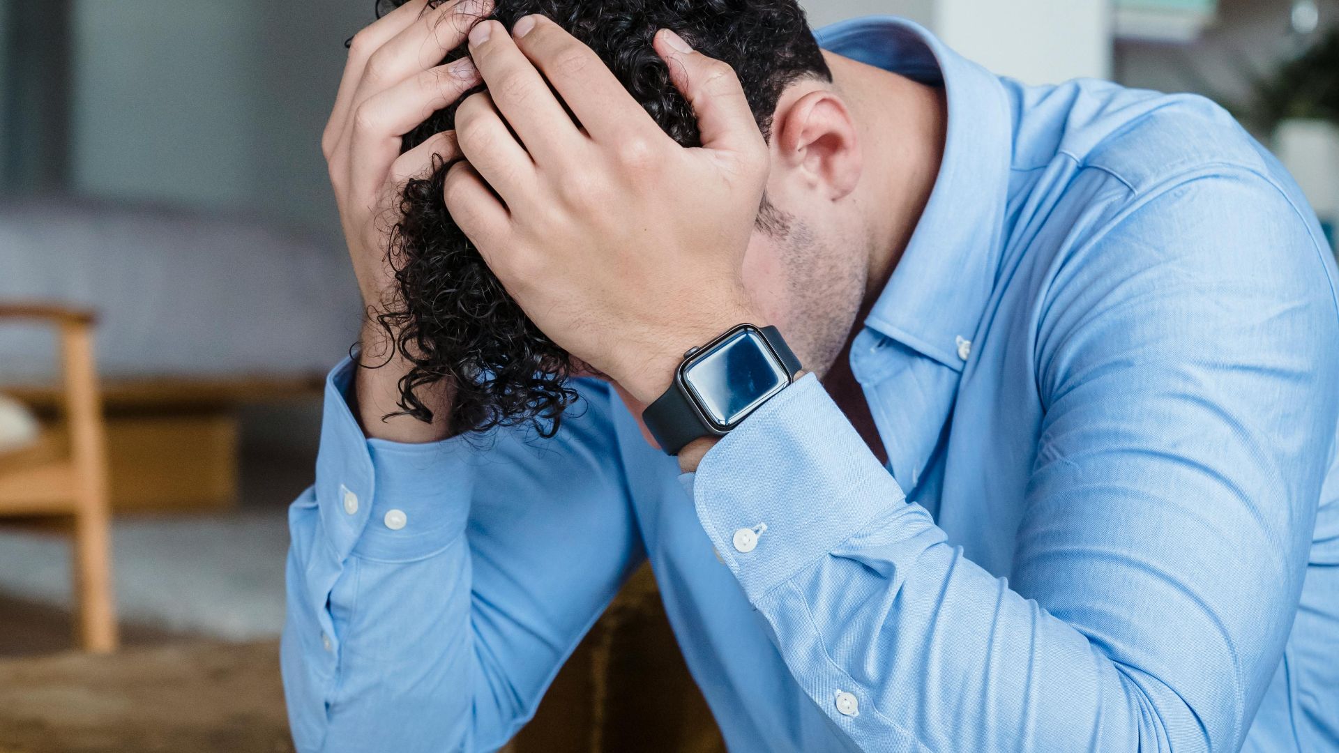 A person showing signs of stress, sitting with hands on head indoors wearing a blue shirt.
