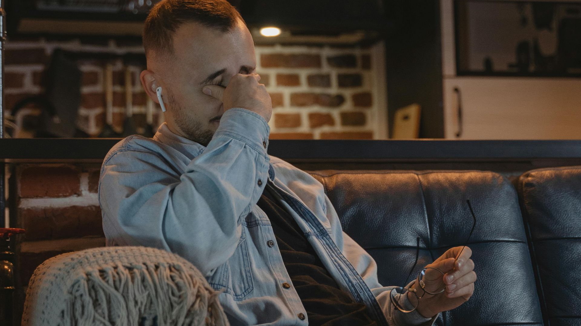 A bearded man in a denim jacket sits on a couch indoors, appearing tired while using a laptop.