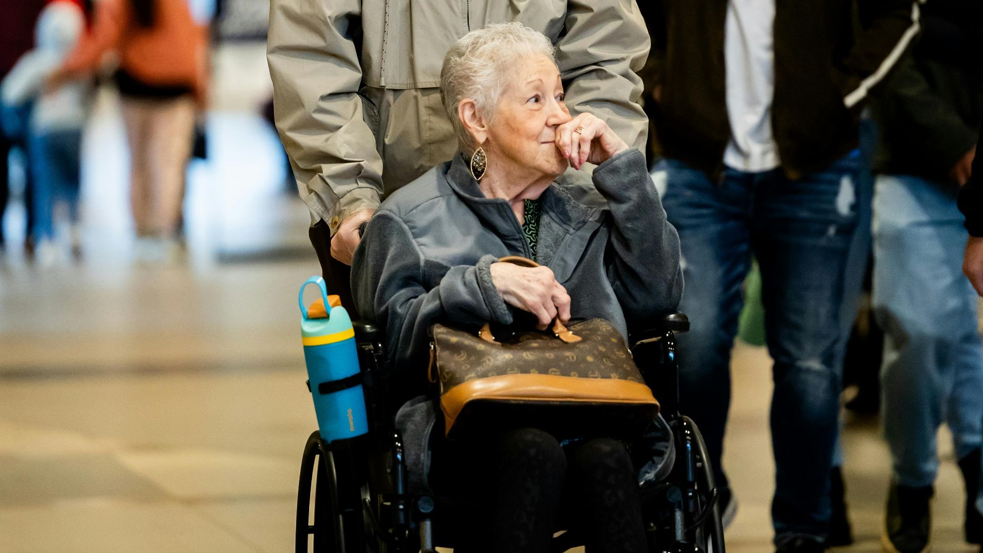 Senior woman in a wheelchair being assisted by a man in a bustling shopping mall.