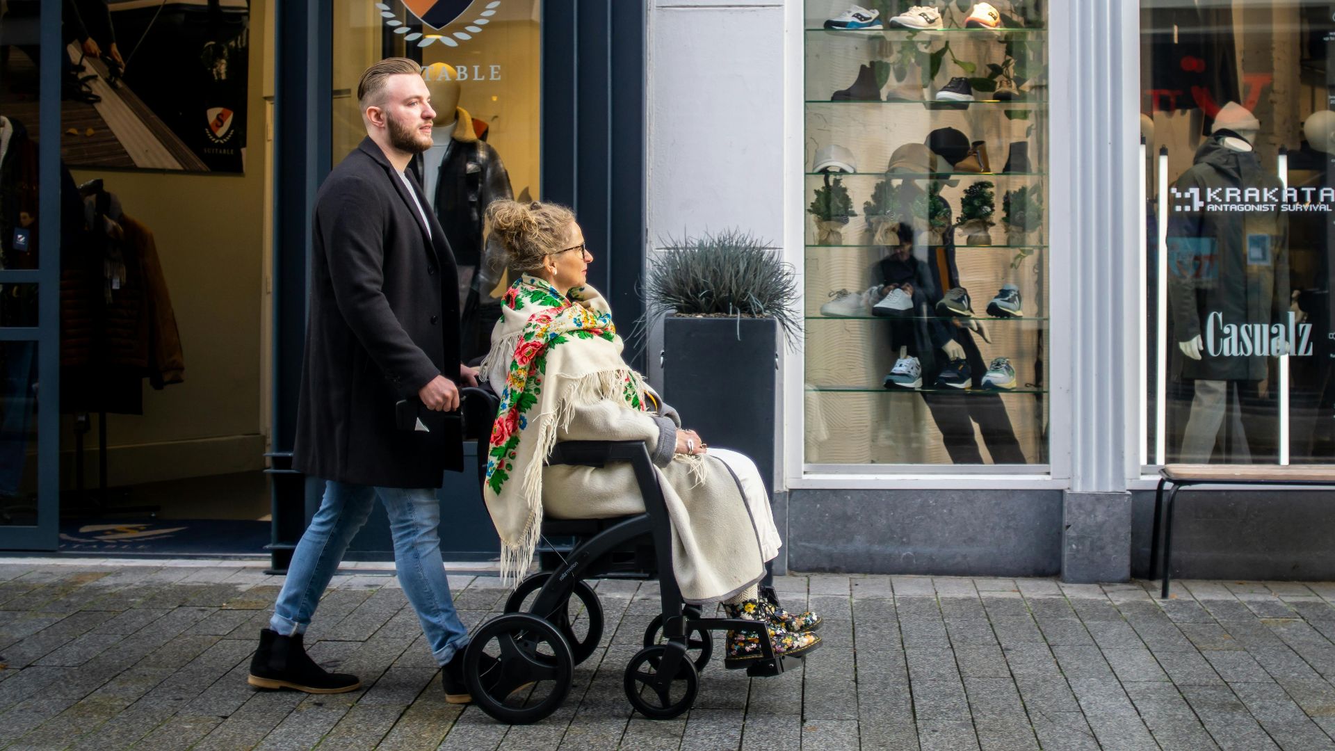 A man helps a woman in a wheelchair outside urban clothing shops, showcasing care and accessibility.