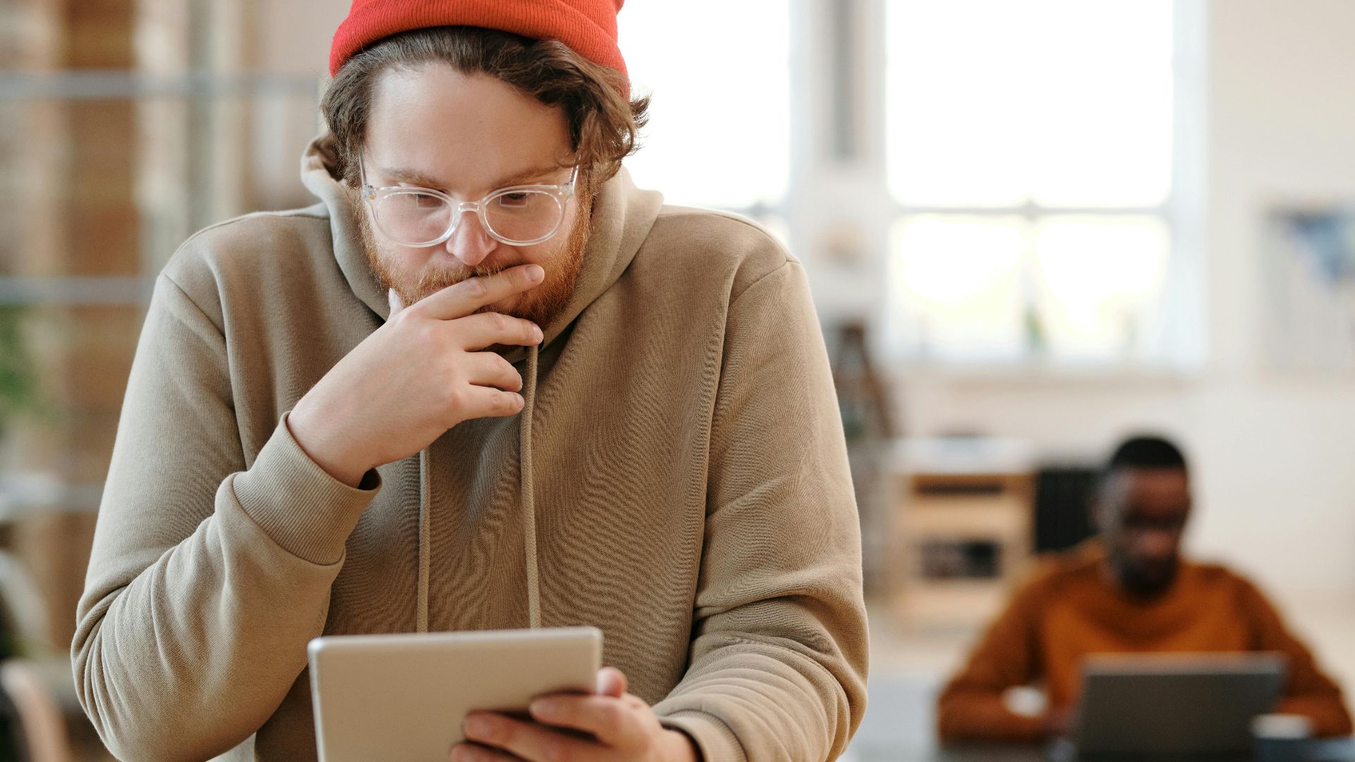 Man in a beanie and glasses using tablet in a bright, modern office.