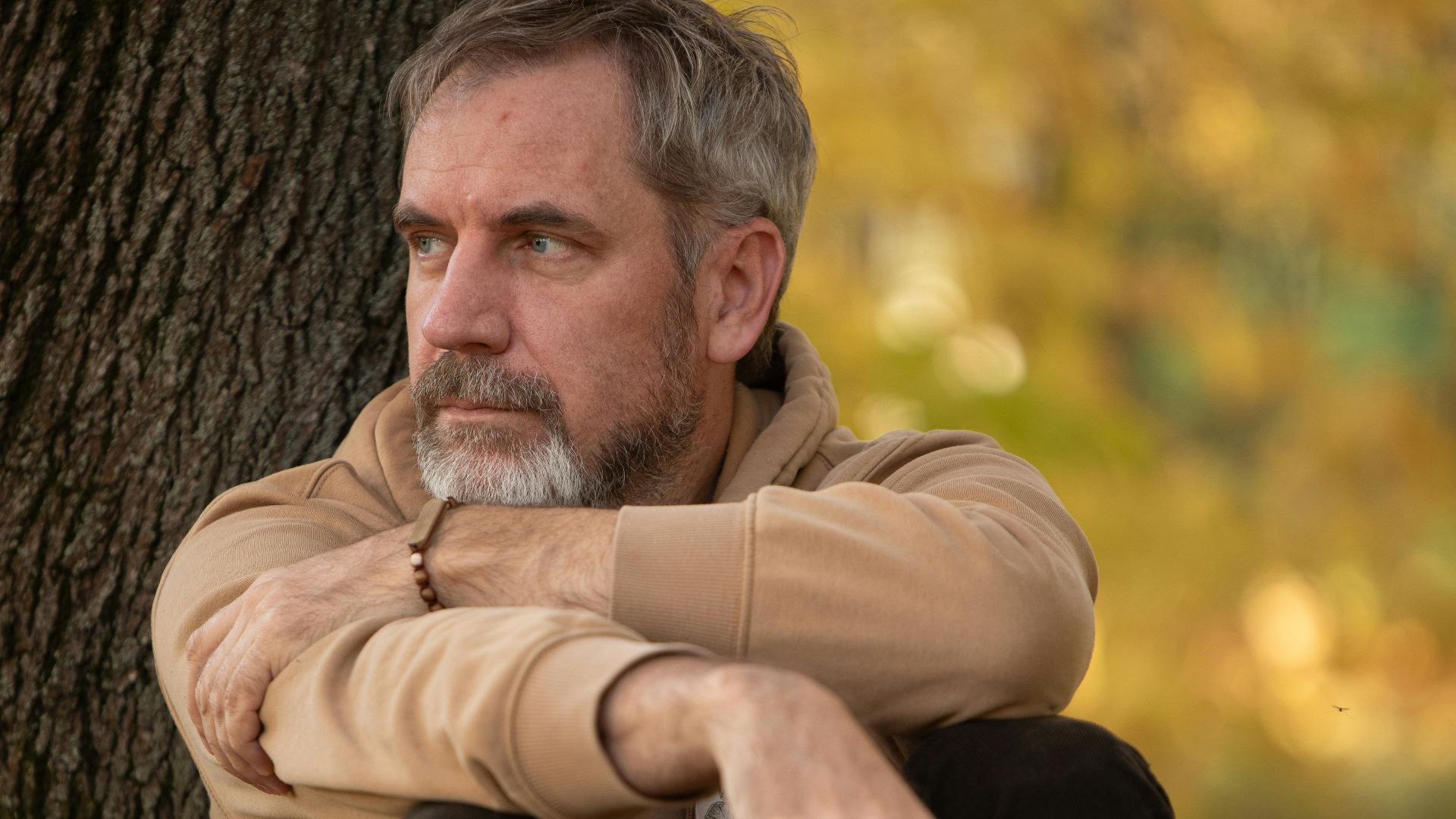 Pensive adult man sitting by a tree in autumn park, deep in thought, serene setting.