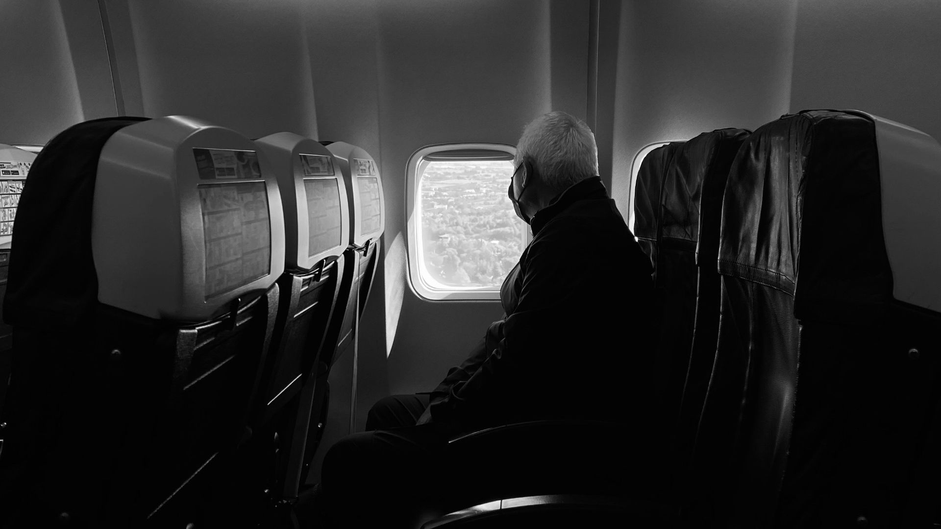 A man wearing a mask gazes out an airplane window during a daytime flight.
