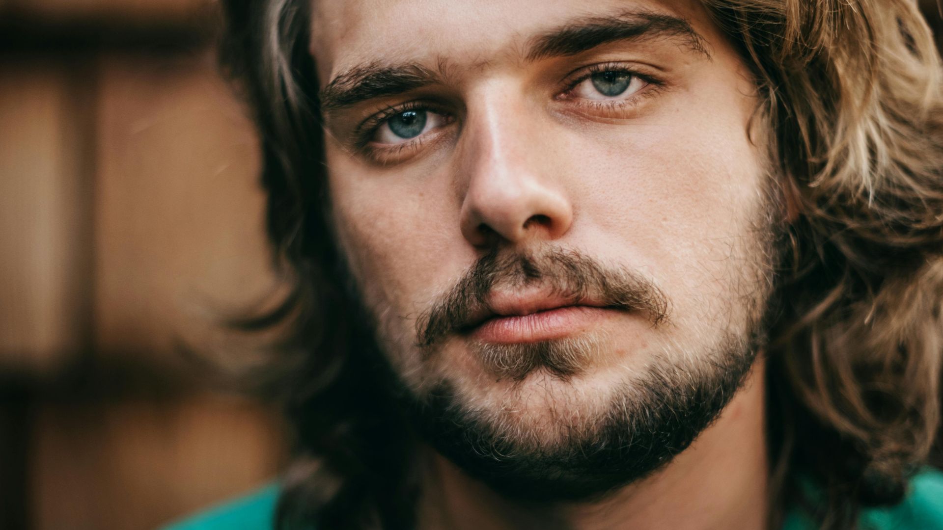 A striking close-up portrait of a young bearded man with long hair and blue eyes.