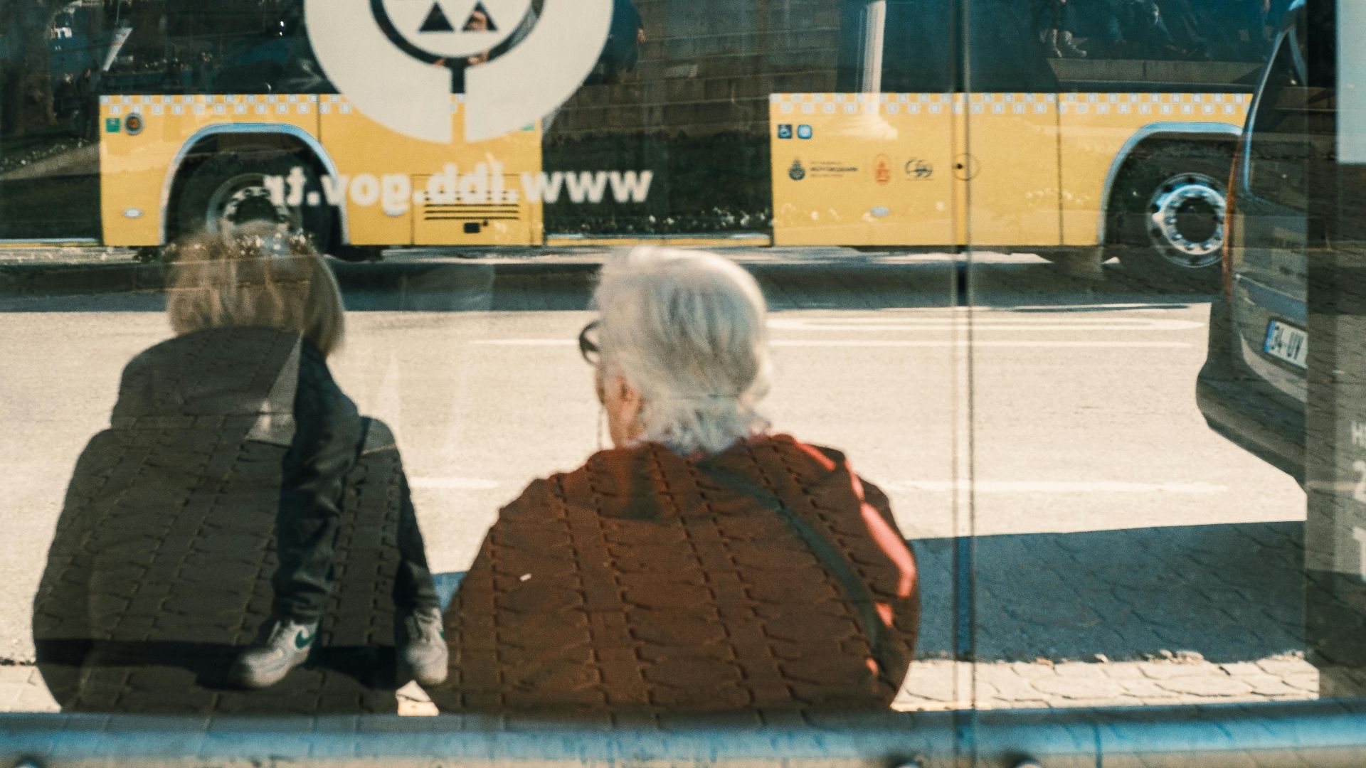 Two individuals at a bus stop with a yellow bus reflection creating an urban scene.