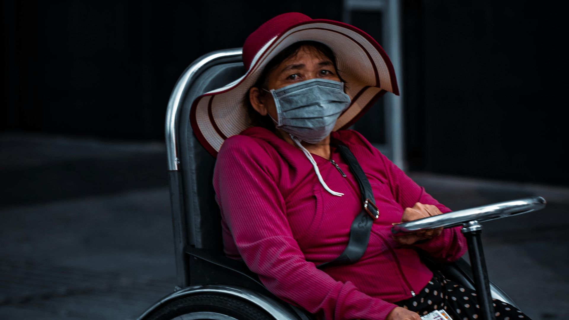 Elderly woman in a wheelchair outdoors, wearing a mask and hat, amid city streets.