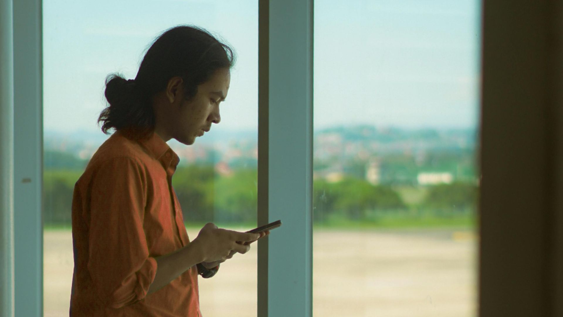 Man in orange shirt using a smartphone while standing at an airport window, overlooking the runway.