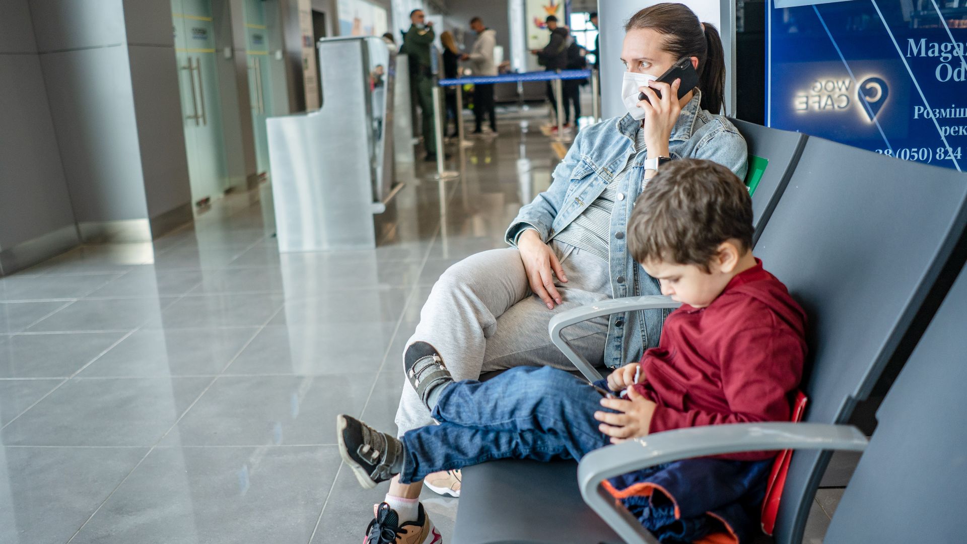 A mother and child sitting at an airport terminal. The mother is on the phone while the child is using a device.
