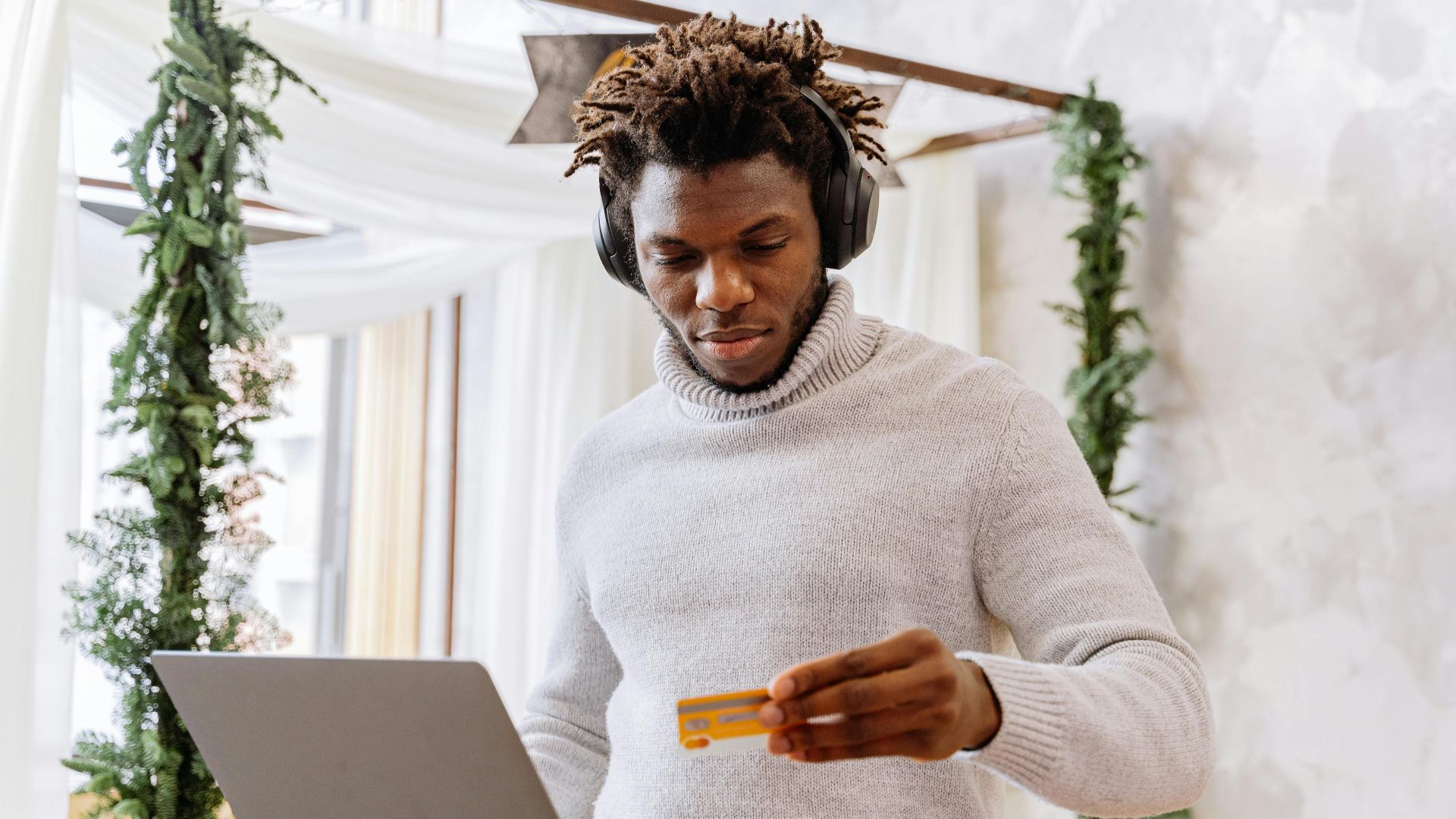 A man stands indoors with a credit card and laptop, wearing headphones and a turtleneck sweater.