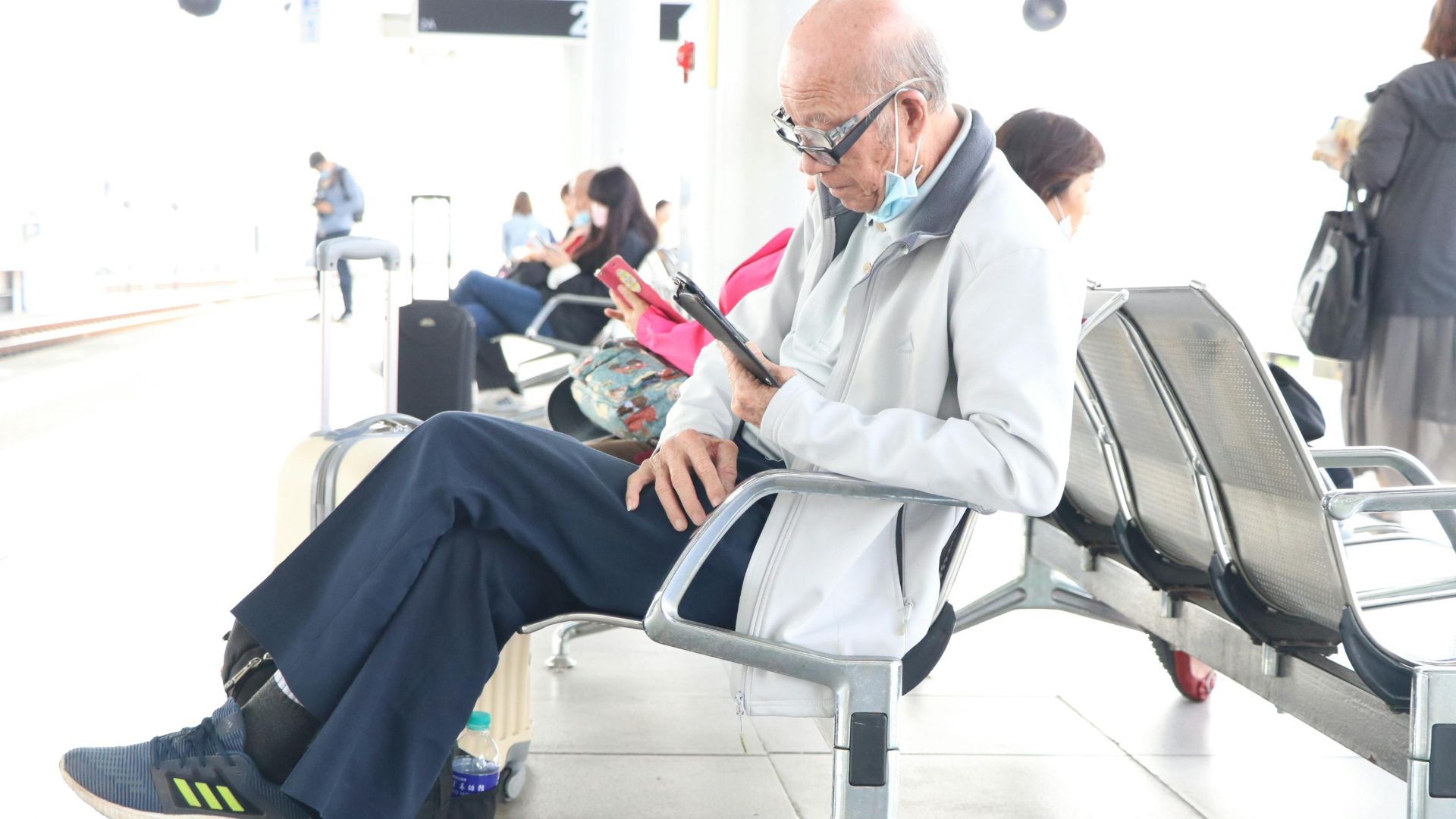 Senior adult using a smartphone in an airport terminal, sitting with luggage nearby.