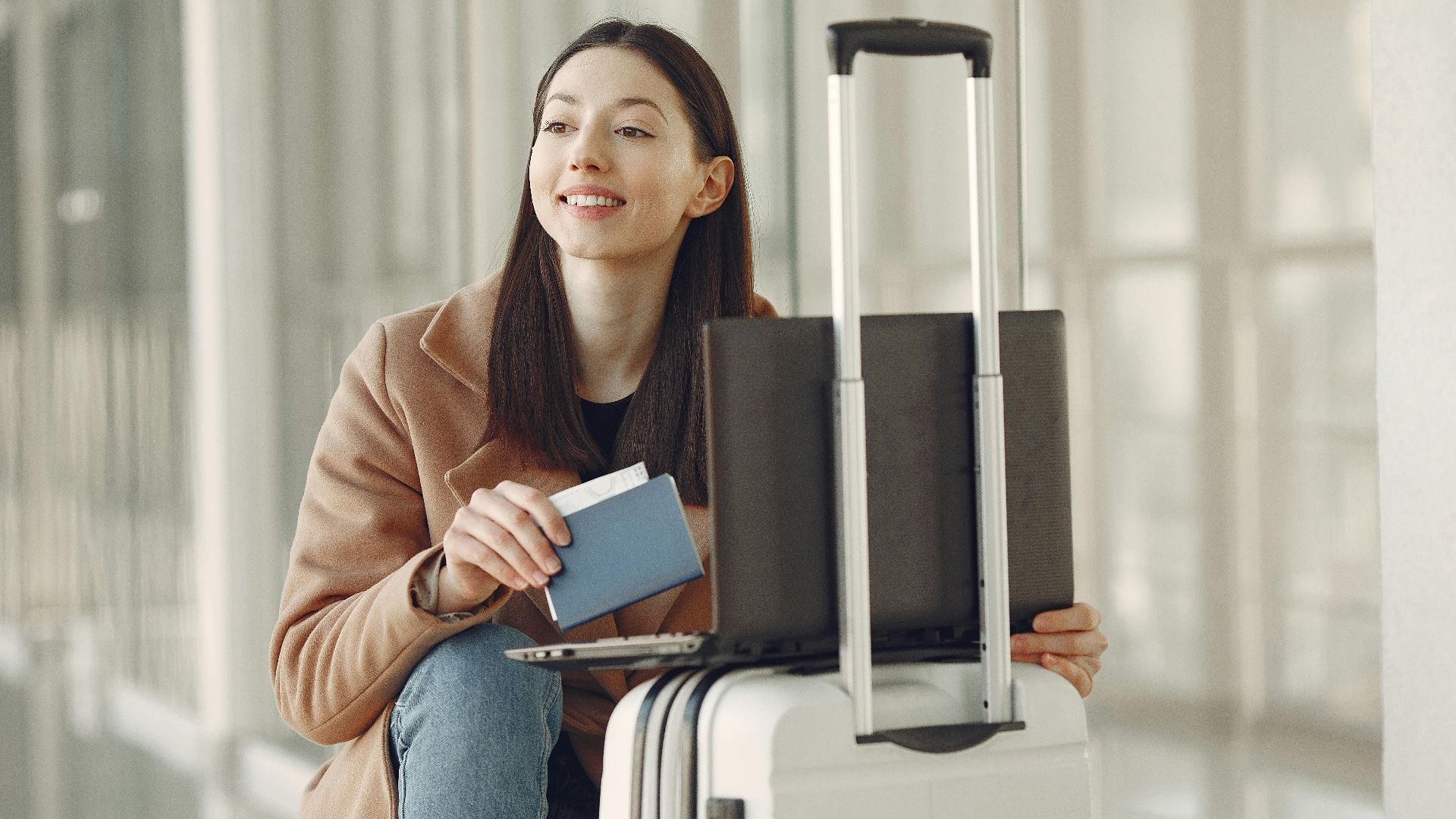 Smiling adult ethnic female traveler in trendy coat holding passport and using laptop on luggage in airport corridor for checking ticket for correctness