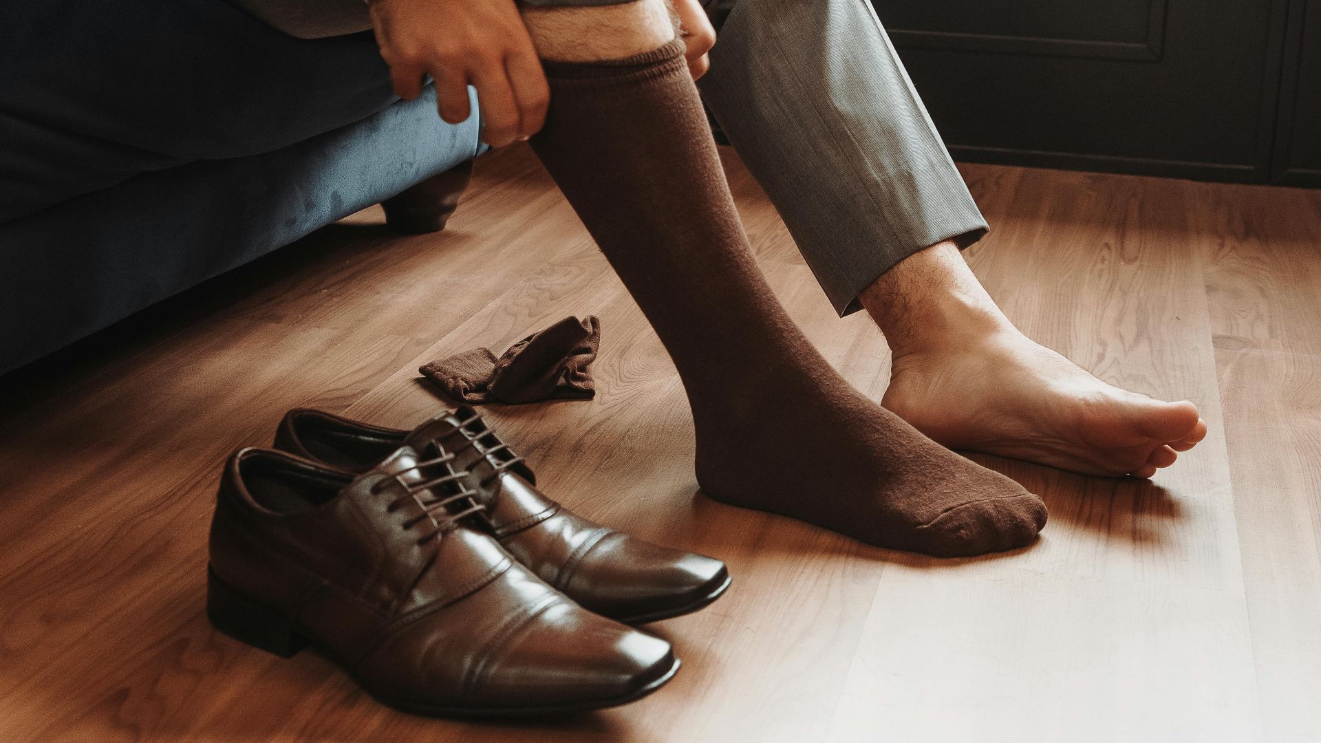 Close-up of a man putting on socks beside elegant brown shoes on a wooden floor.