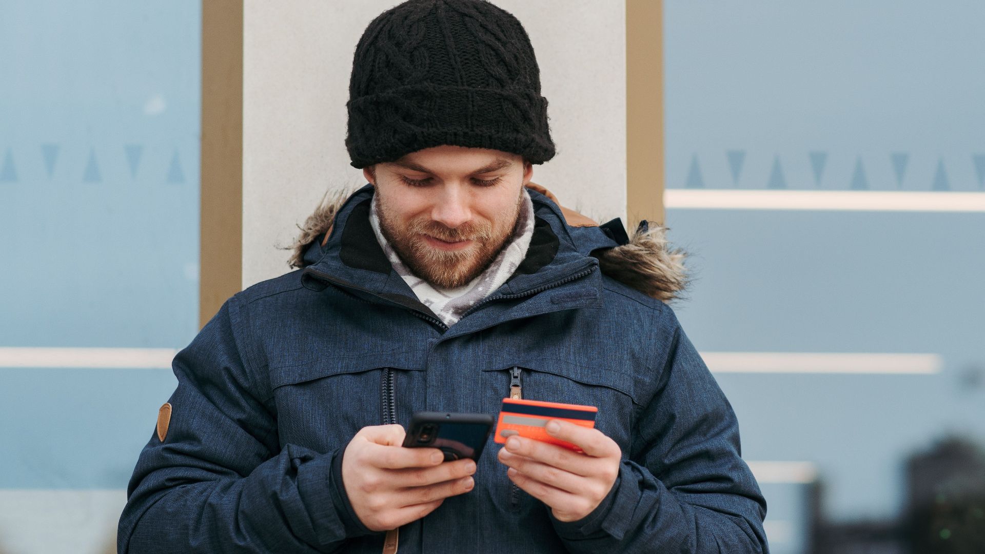 Positive man in warm clothes standing near building and entering credentials of credit card on mobile phone in daylight