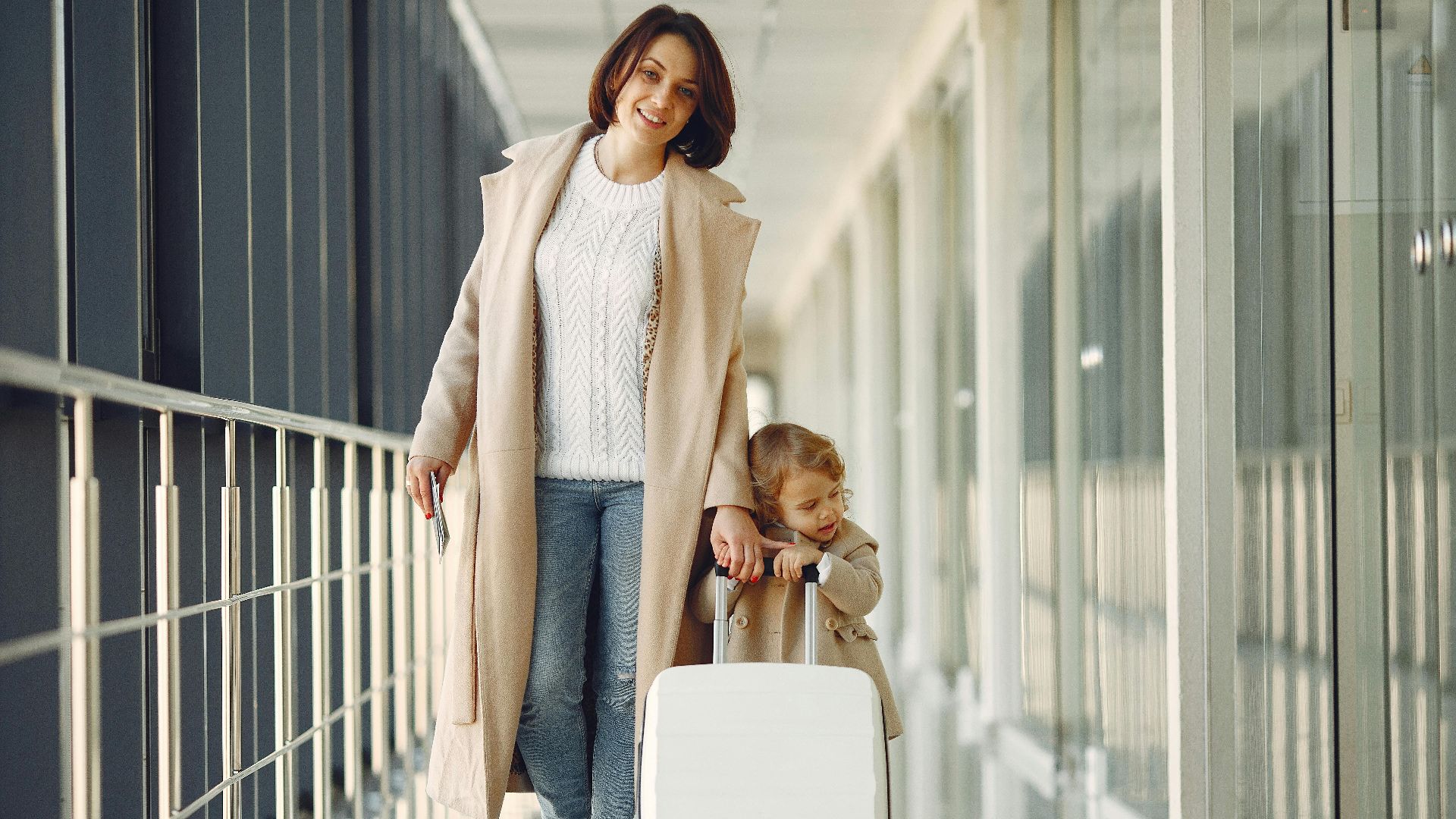 Full length of content woman with daughter in jeans and beige coats carrying white suitcase while walking along modern airport airbridge