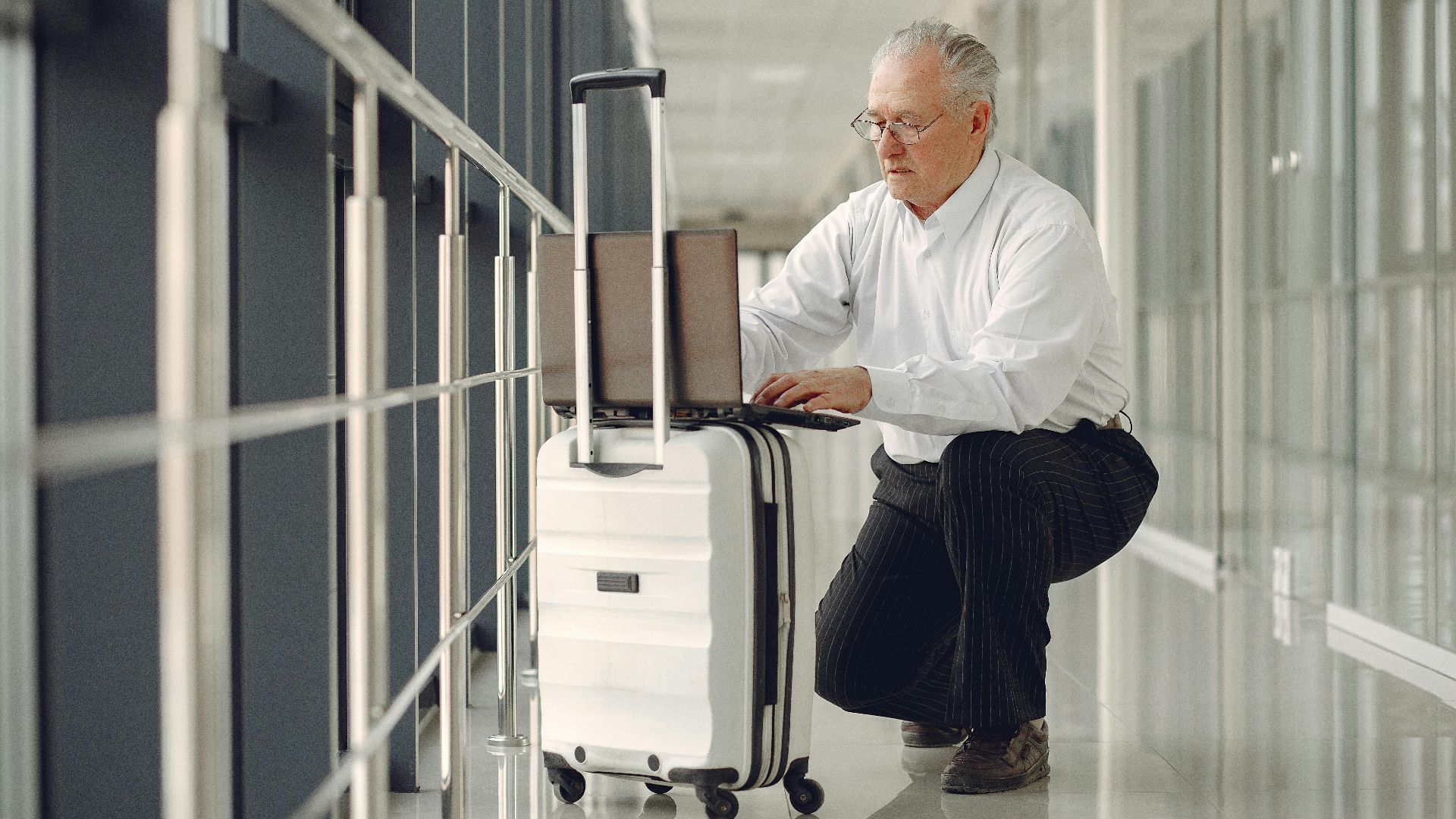 Full length of focused mature male in formal wear using laptop on baggage while having semisitting position in airport corridor