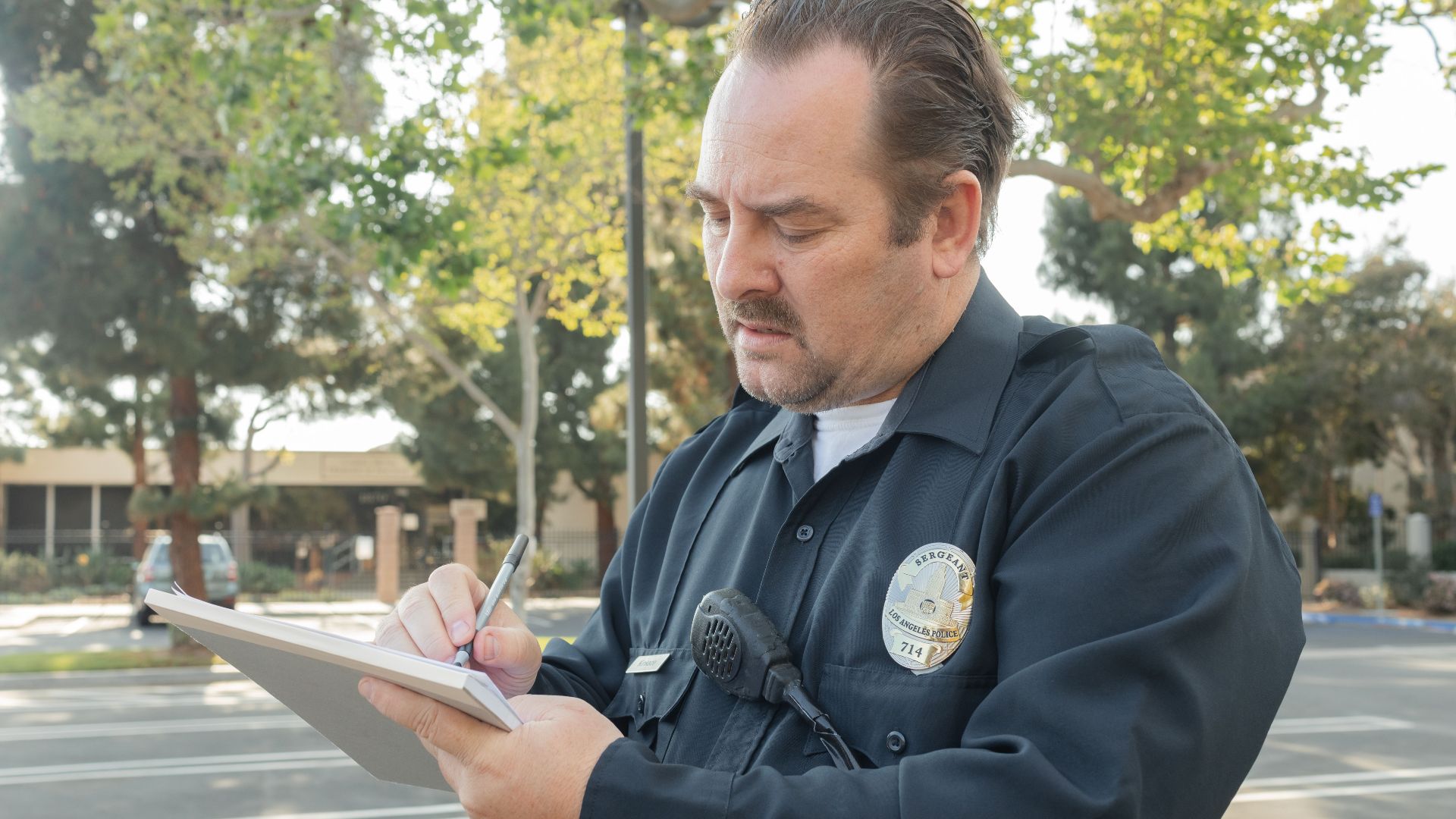 A police officer outdoors writing a report on a notepad. Trees in the background.