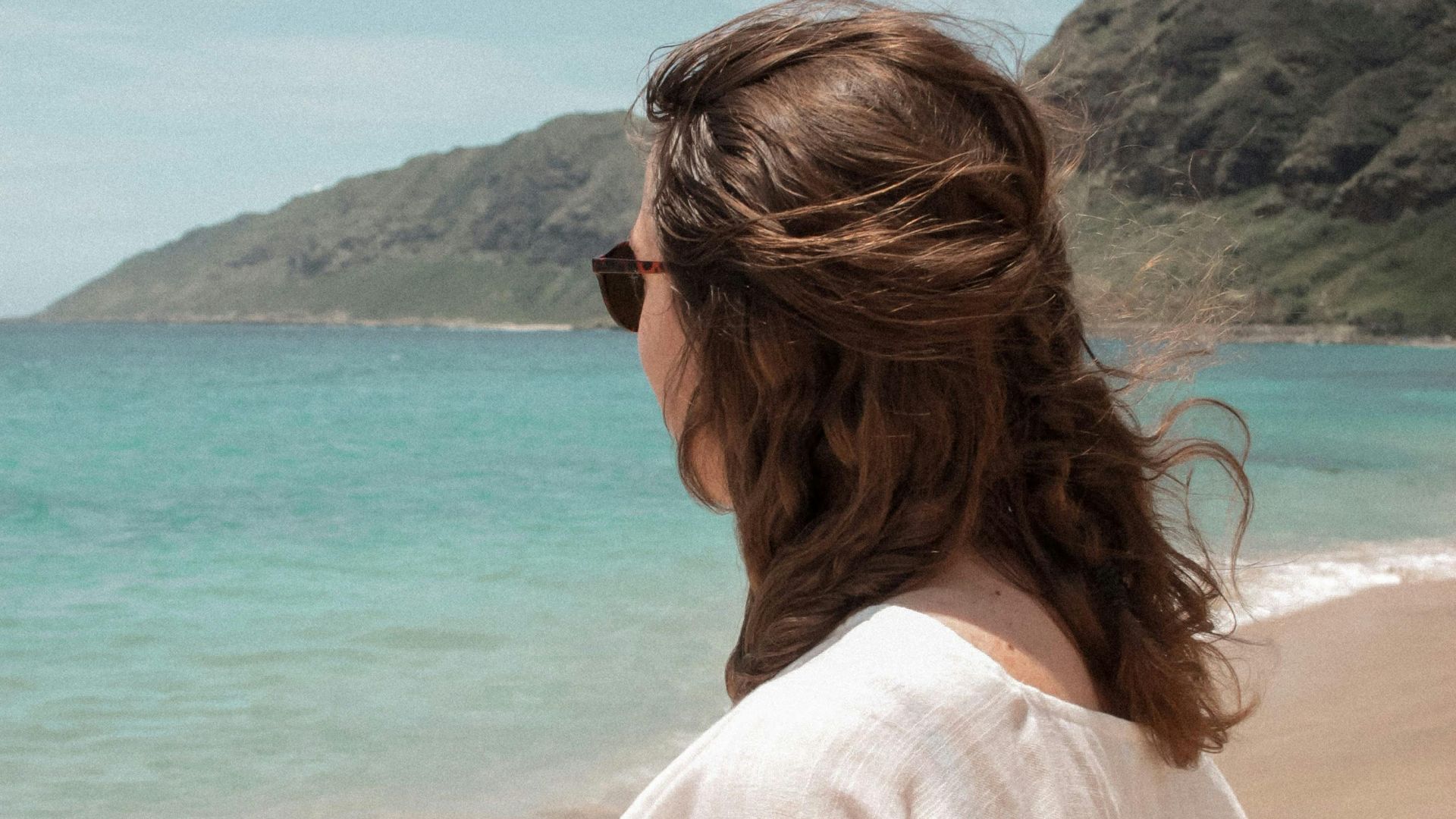 Woman in white outfit enjoying a sunny day at the beach with mountains in the distance.