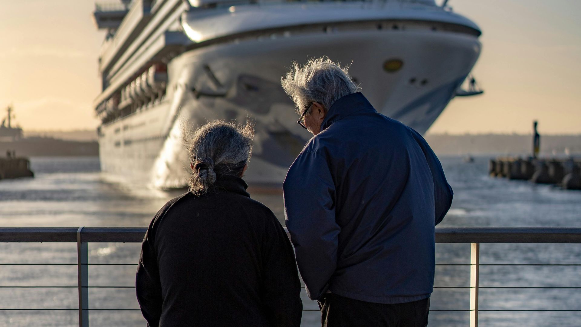 Senior couple standing at railing, admiring large cruise ship approaching dock at sunset.