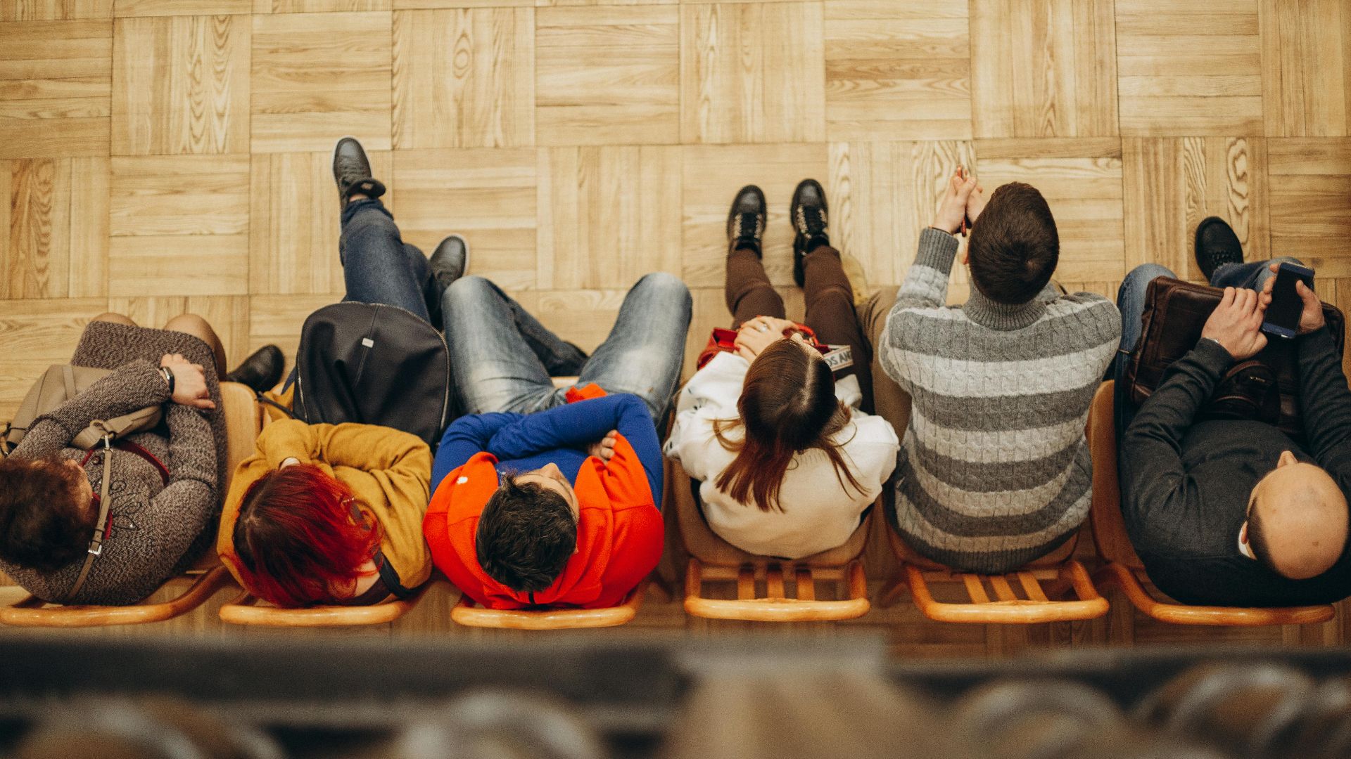 Top view of adults sitting in a row on wooden chairs on a parquet floor indoors.