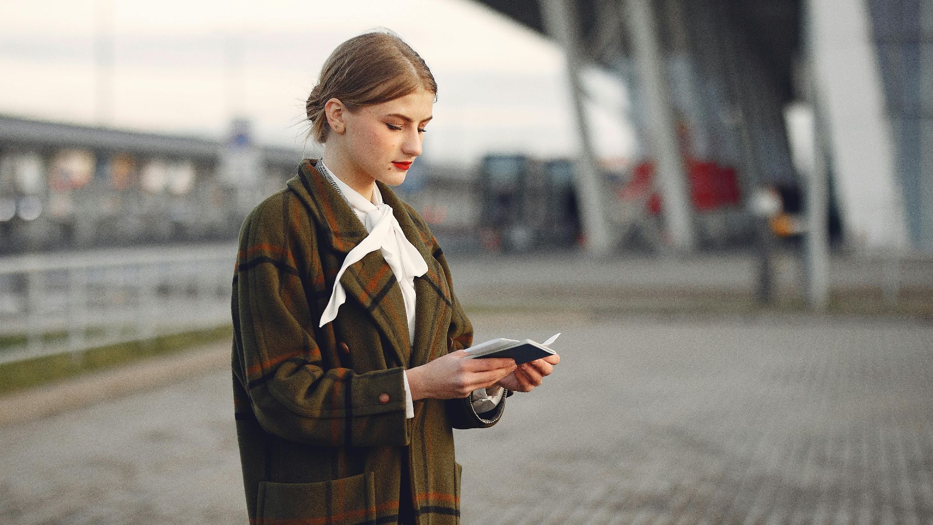 Attentive female passenger wearing trendy plaid coat and white blouse checking passport and ticket standing on pavement near modern building of airport outside