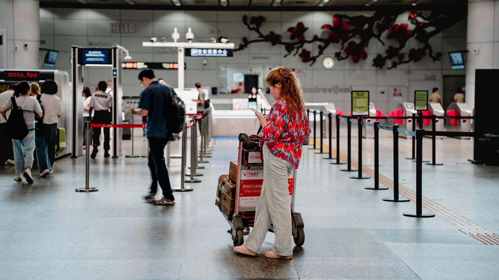 A traveler checks her phone while waiting with a luggage cart at a busy airport terminal in Guangzhou.