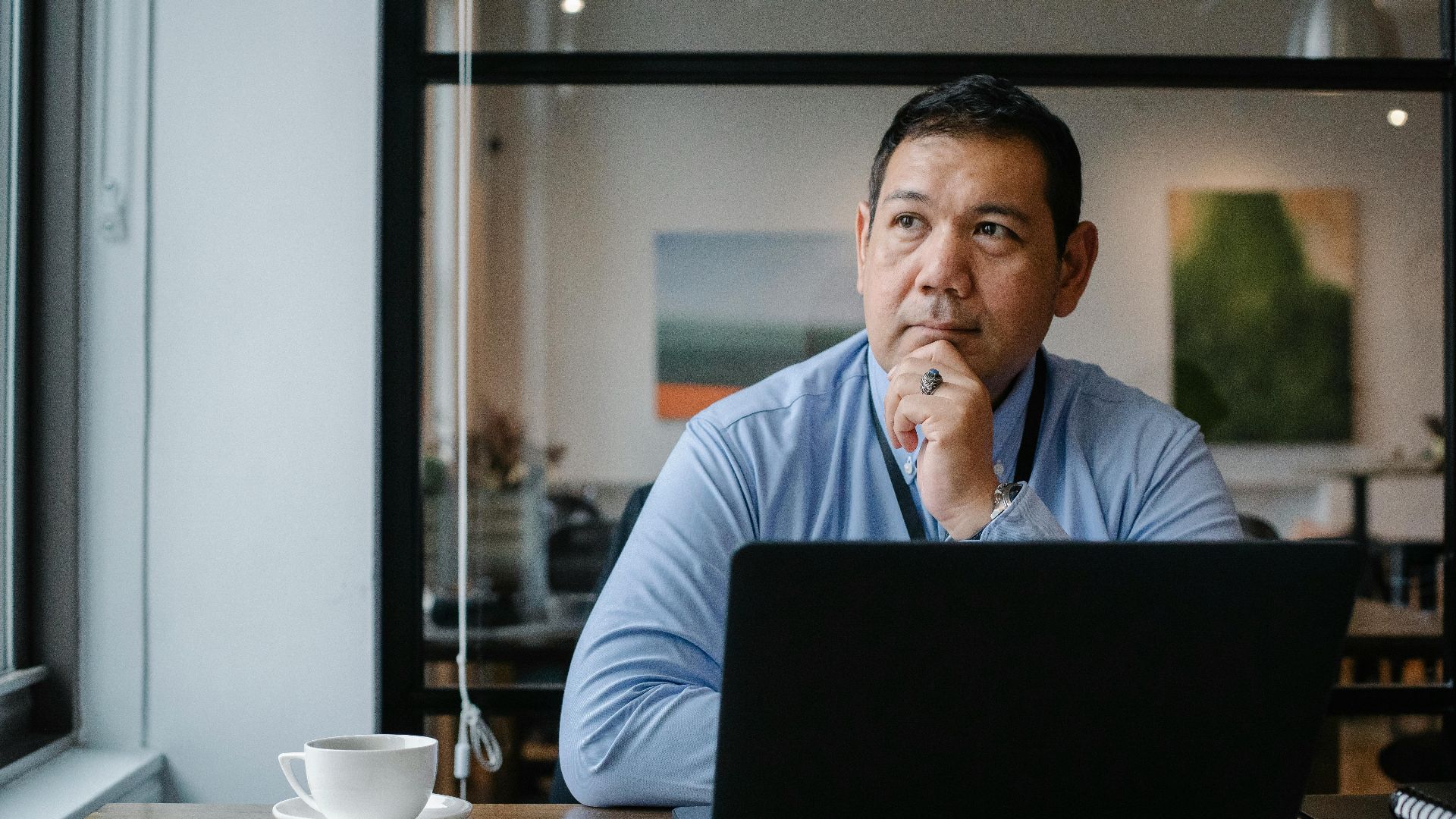 Concentrated ethnic adult male entrepreneur thinking on project while working at table with netbook and cup of drink