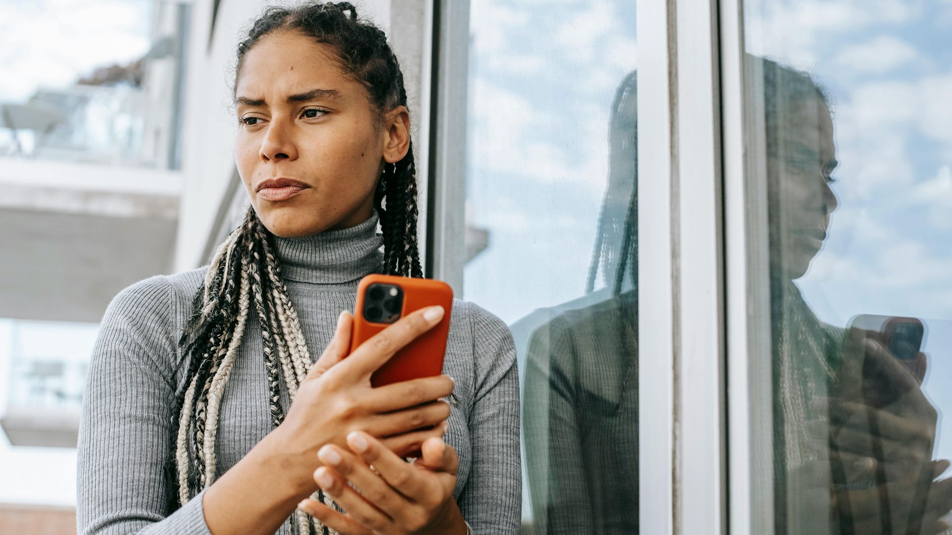 Low angle of concerned ethnic female with smartphone in stressful situation near glass wall
