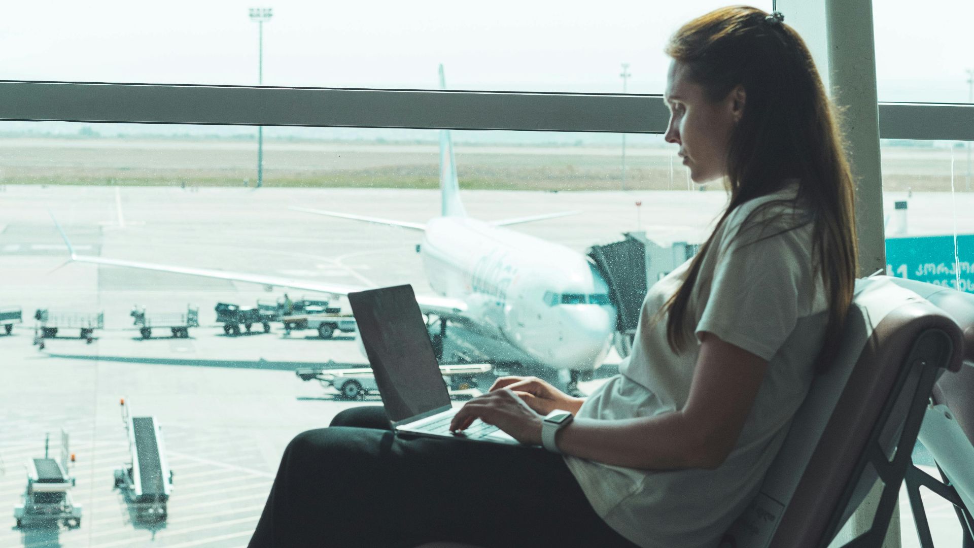 Side view of woman using laptop at airport, airplane visible outside through large glass windows.