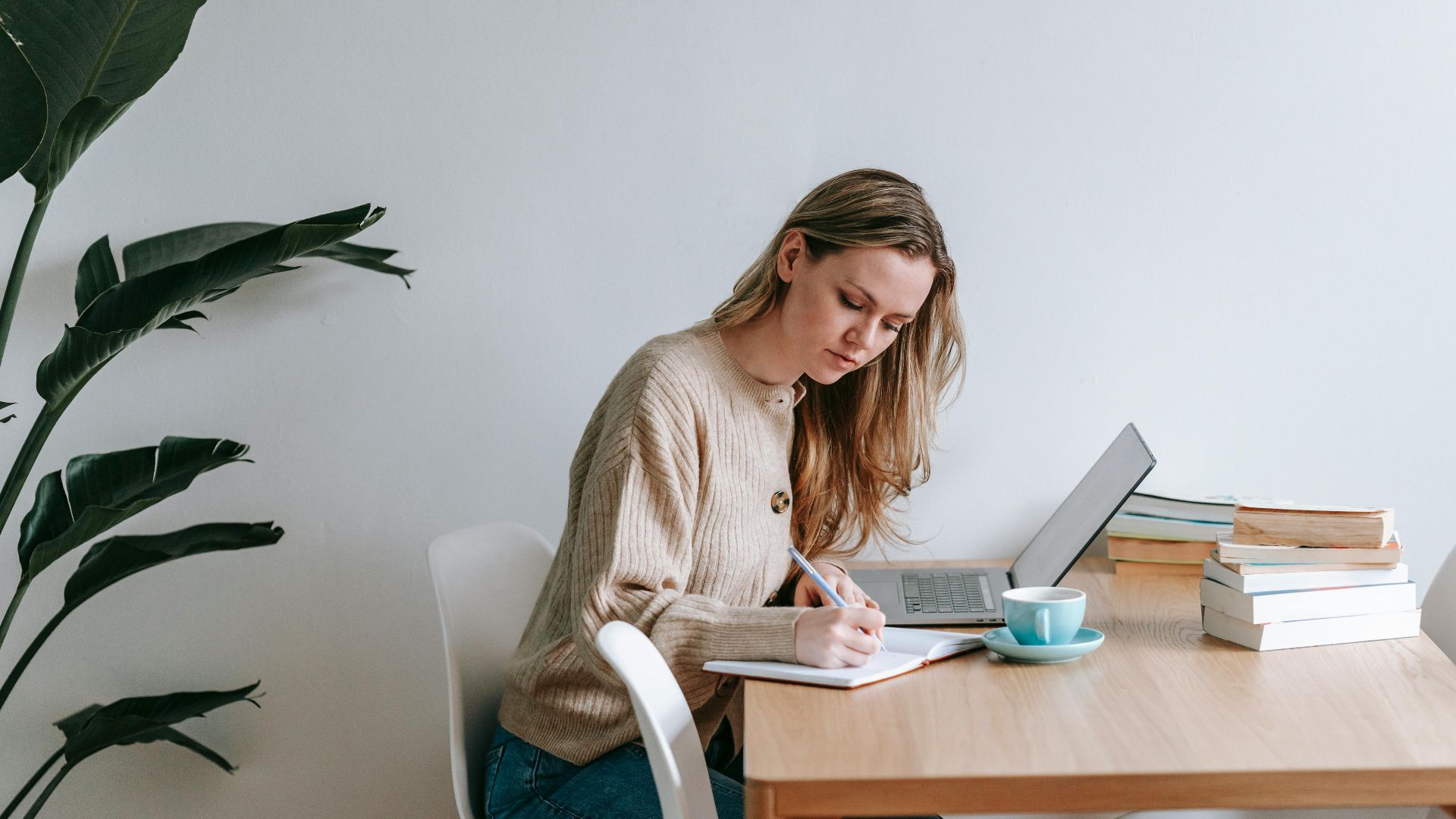 Side view of concentrated female freelancer taking notes in notepad while working at table with netbook in light room with green plant