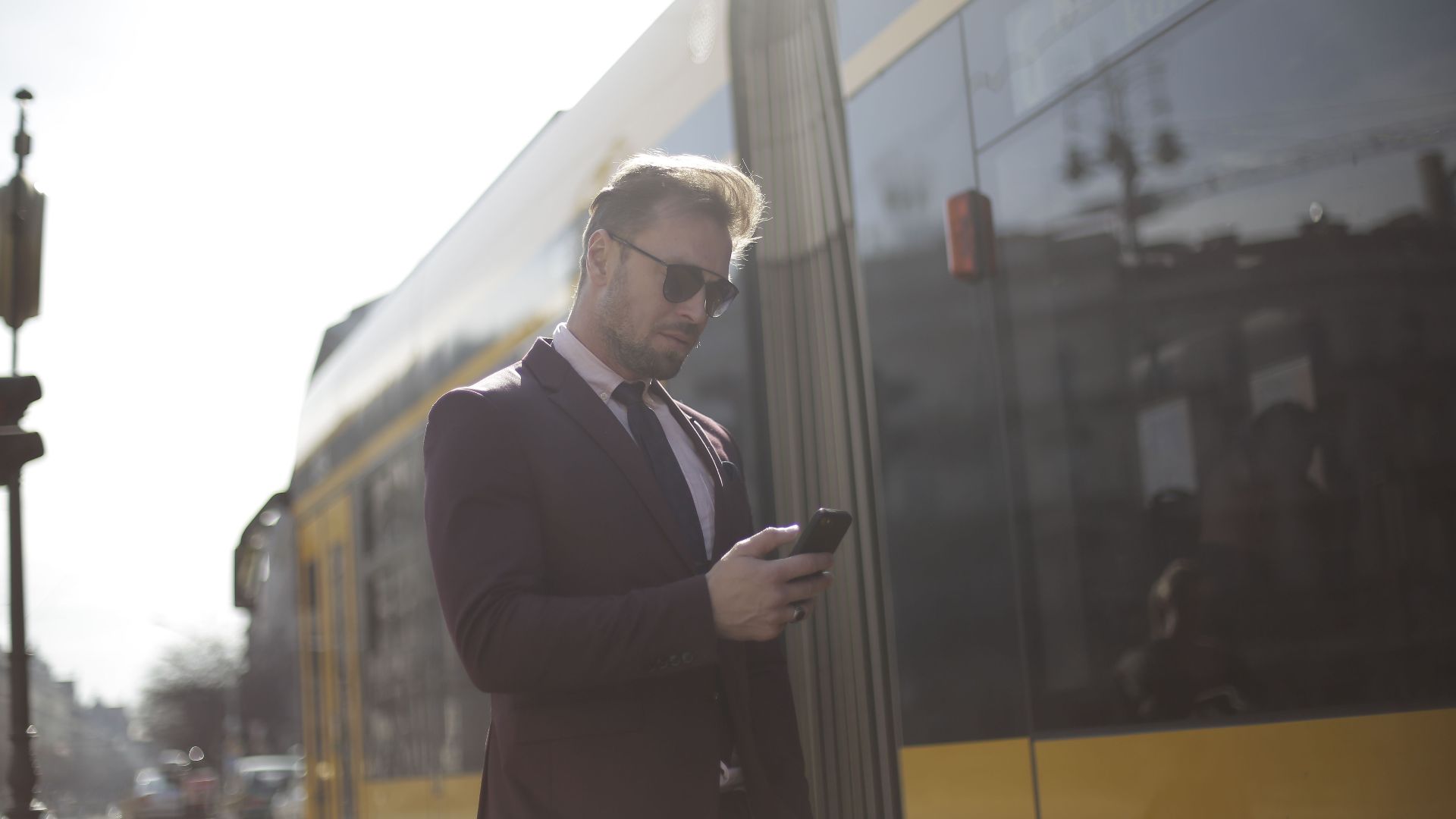 Business professional checks phone at a sunny train station, wearing a suit and sunglasses.