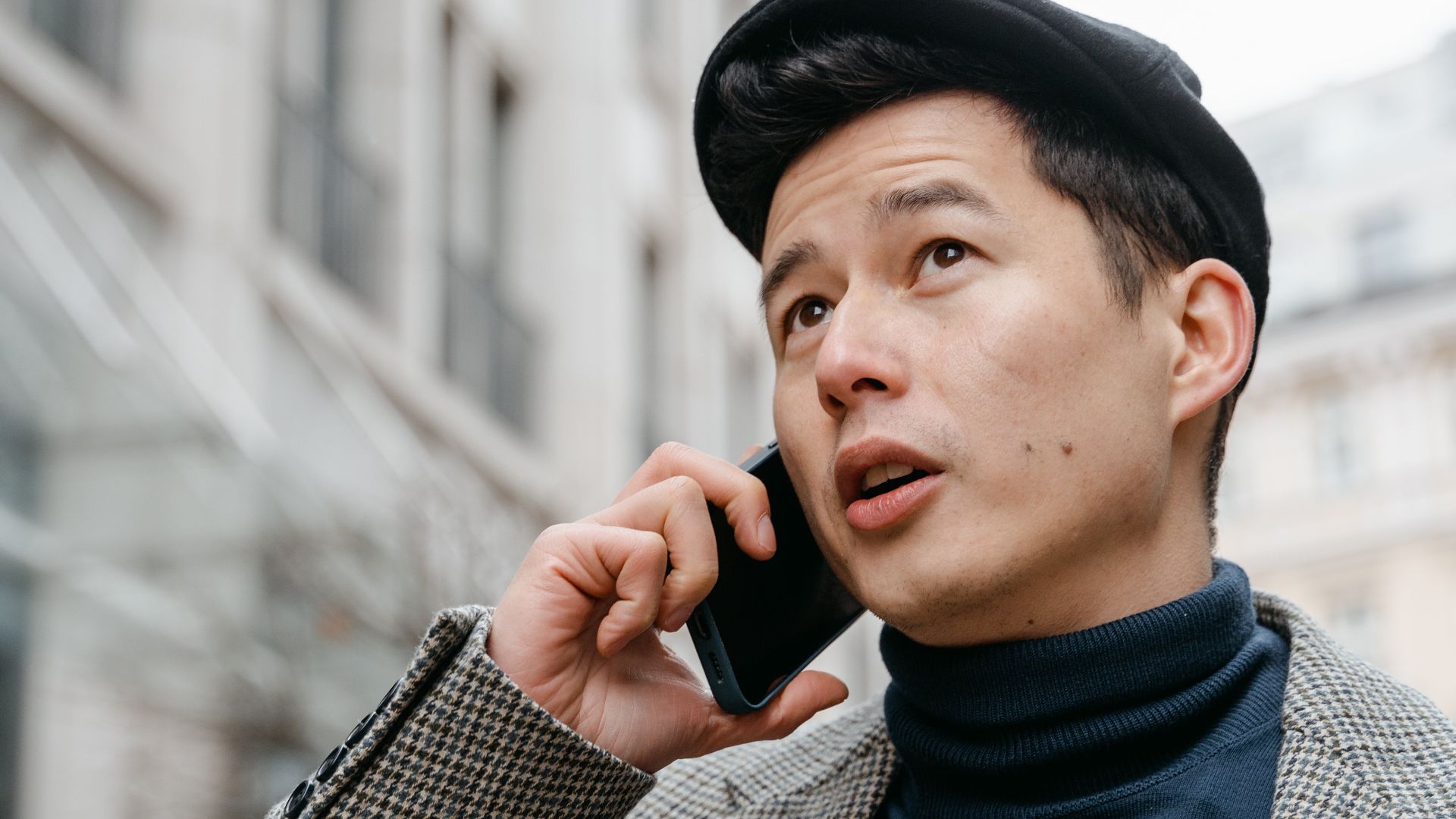 Stylish man in a city talking on a smartphone outdoors, wearing a coat and hat.