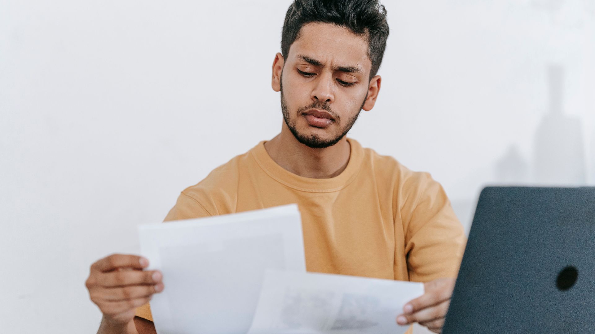 Attentive young bearded Hispanic male remote worker examining papers with results of project while working with laptop in home office