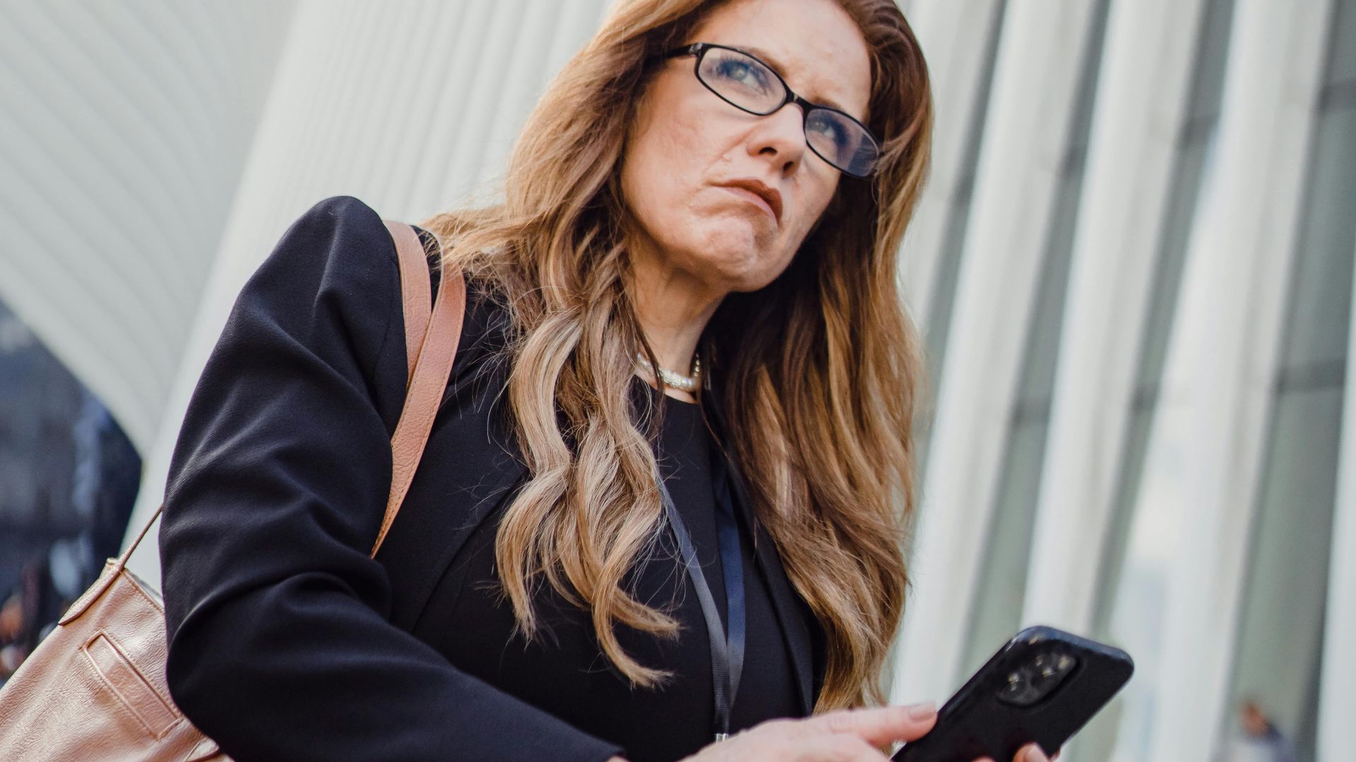 Redhead woman in black suit with brown handbag using smartphone against urban architecture.