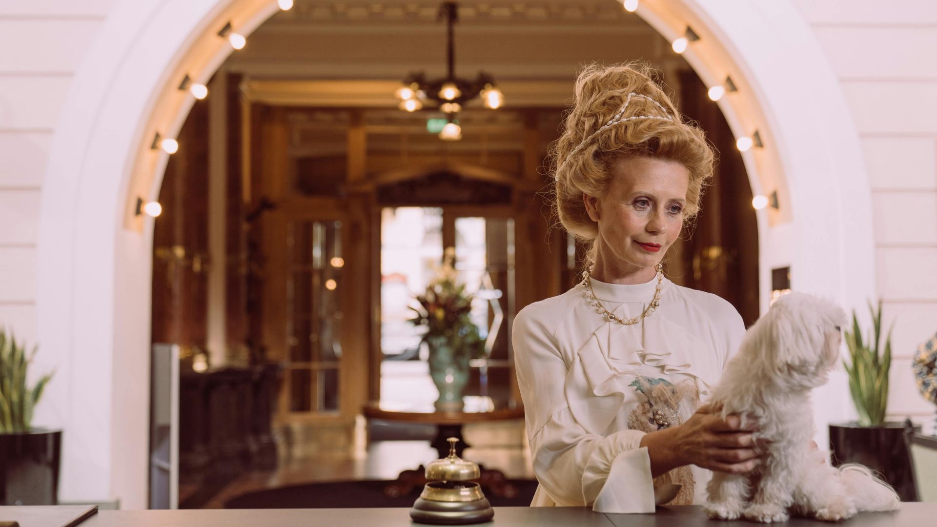 An elegant woman holding a dog at a luxurious hotel reception desk.