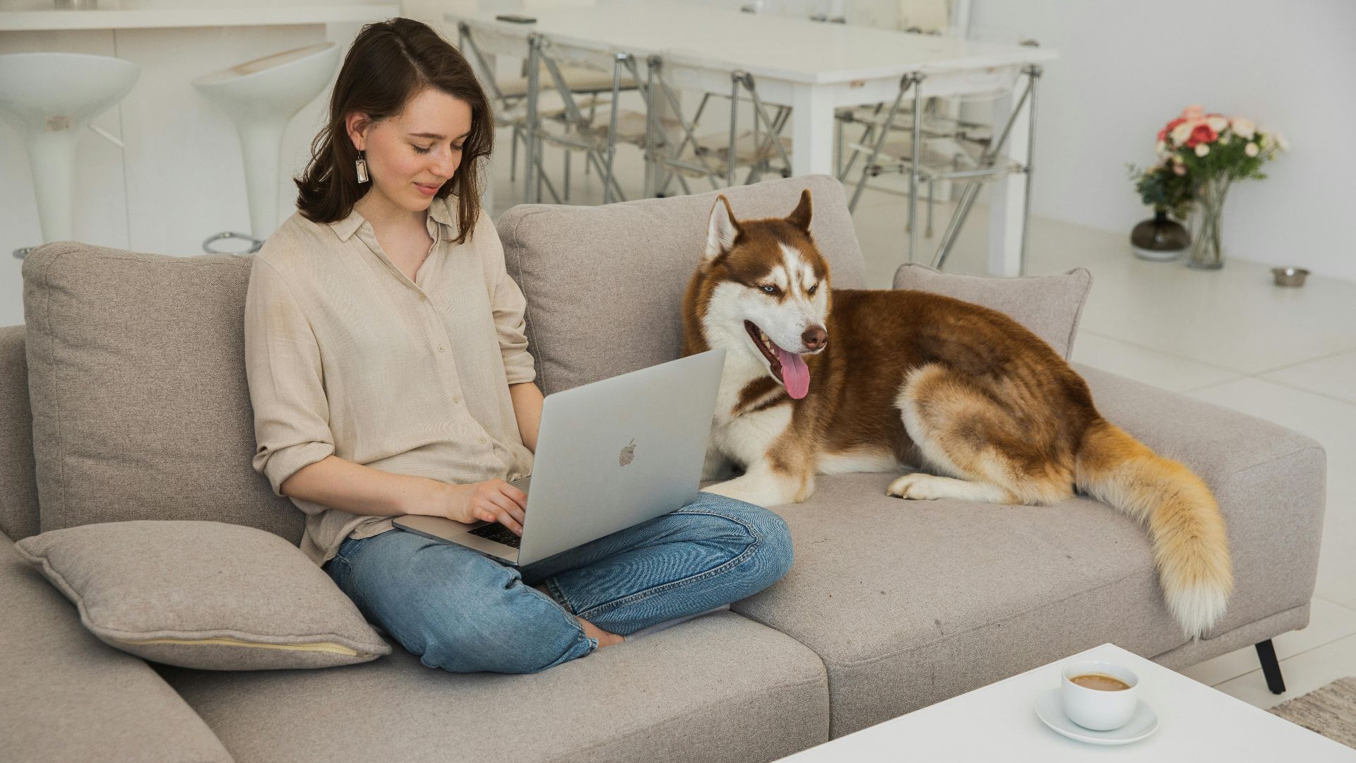 Woman sits with her Husky dog while using laptop, in a cozy living room.