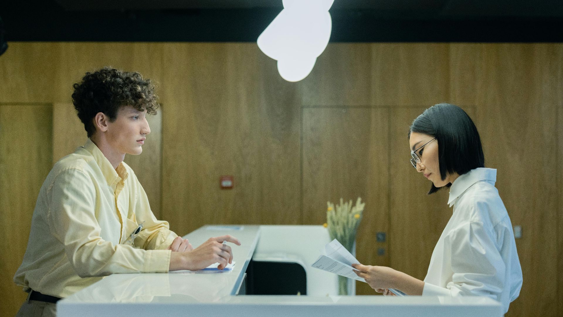 A young man and woman interacting at a modern office reception desk indoors.
