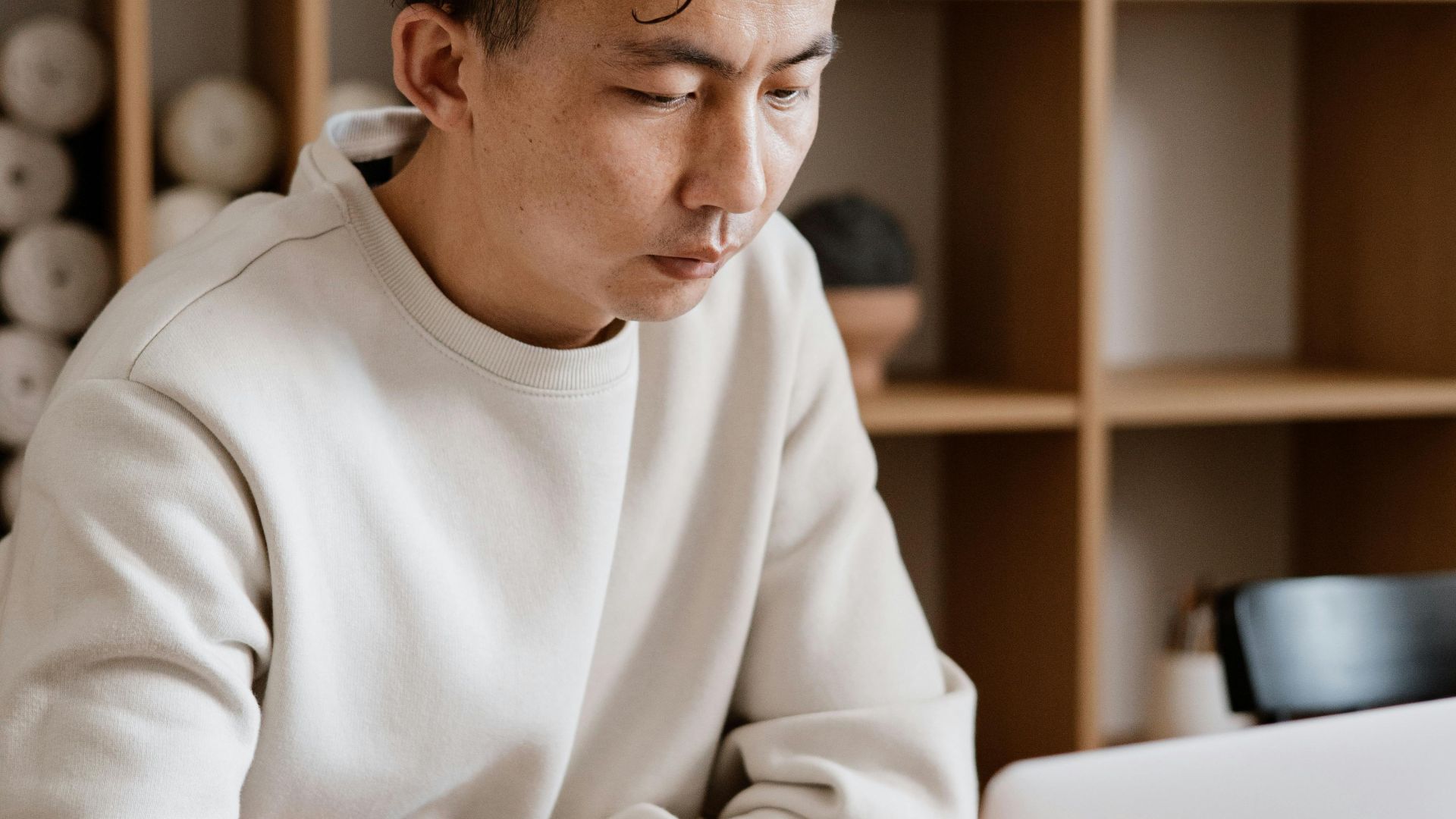 Asian man in a sweater working on a laptop in a modern office setting.