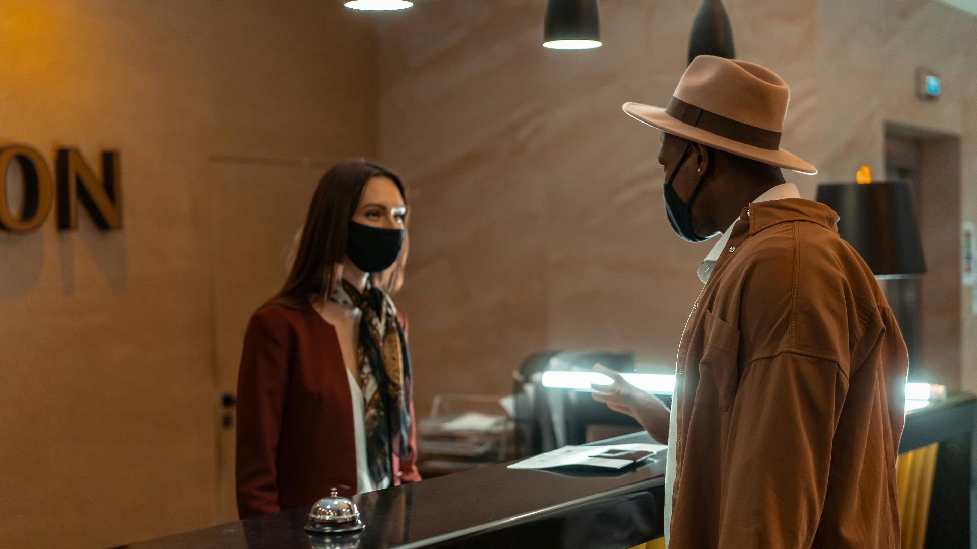 A man at a hotel reception talking to a staff member. Both wear face masks.