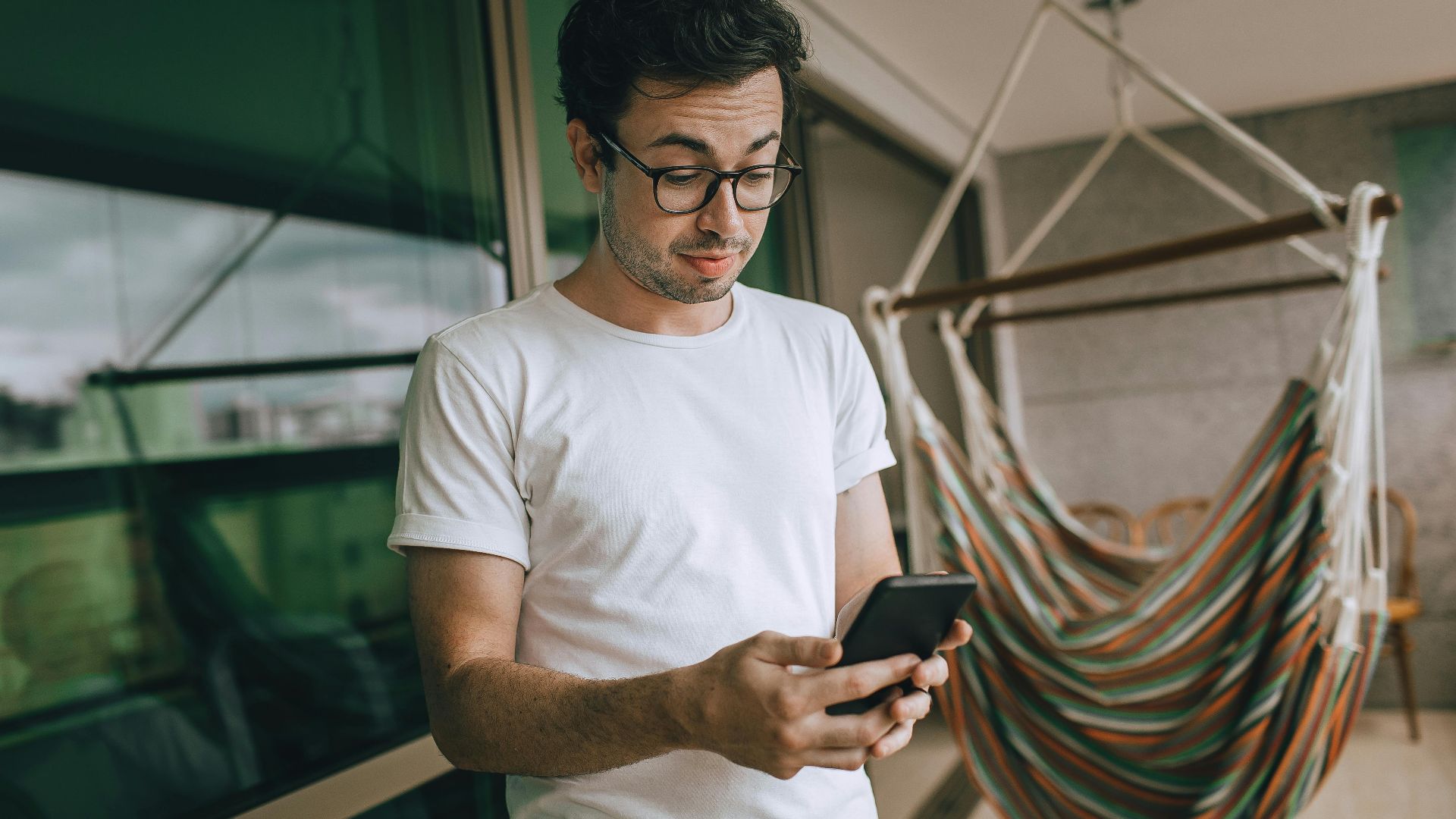 Young man in glasses using smartphone indoors with striped hammock.