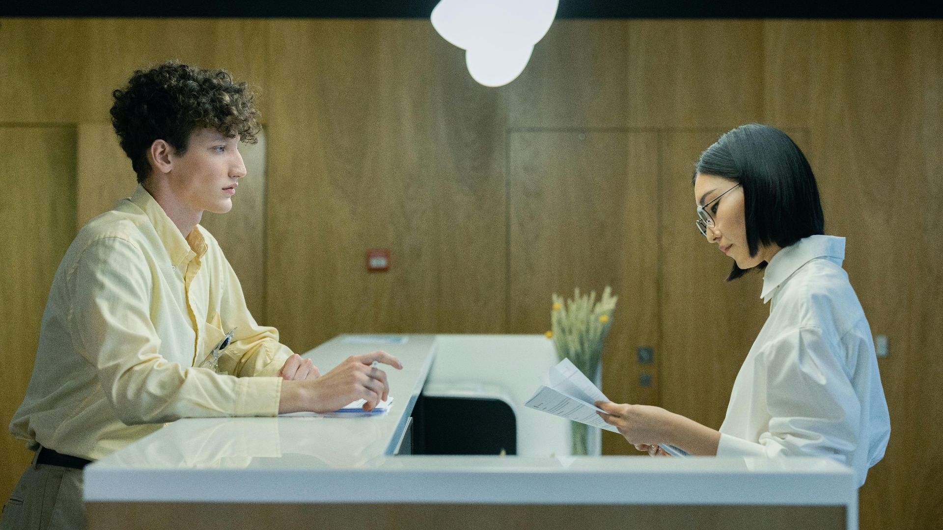 A young man and woman interacting at a modern office reception desk indoors.