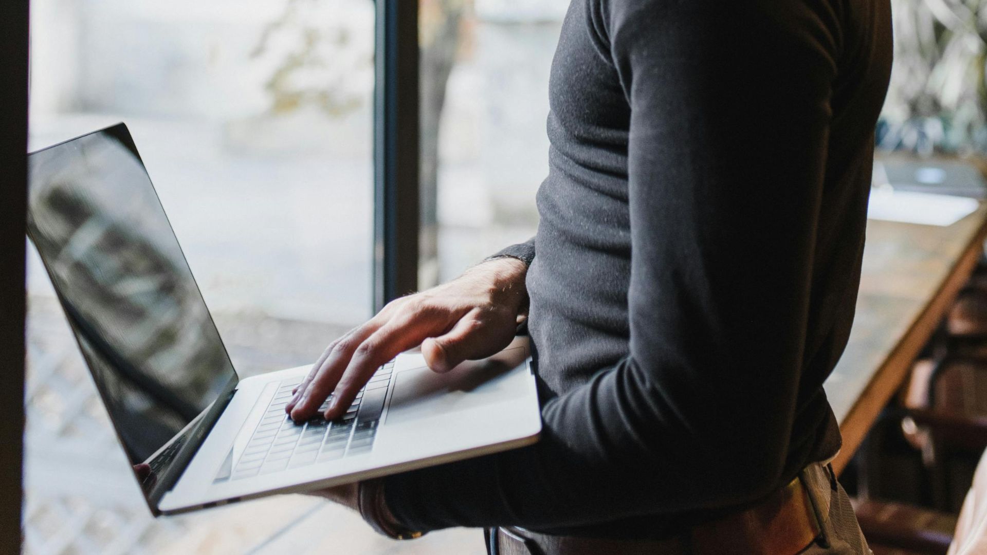 Adult man using a laptop indoors by a large window, focused on work.