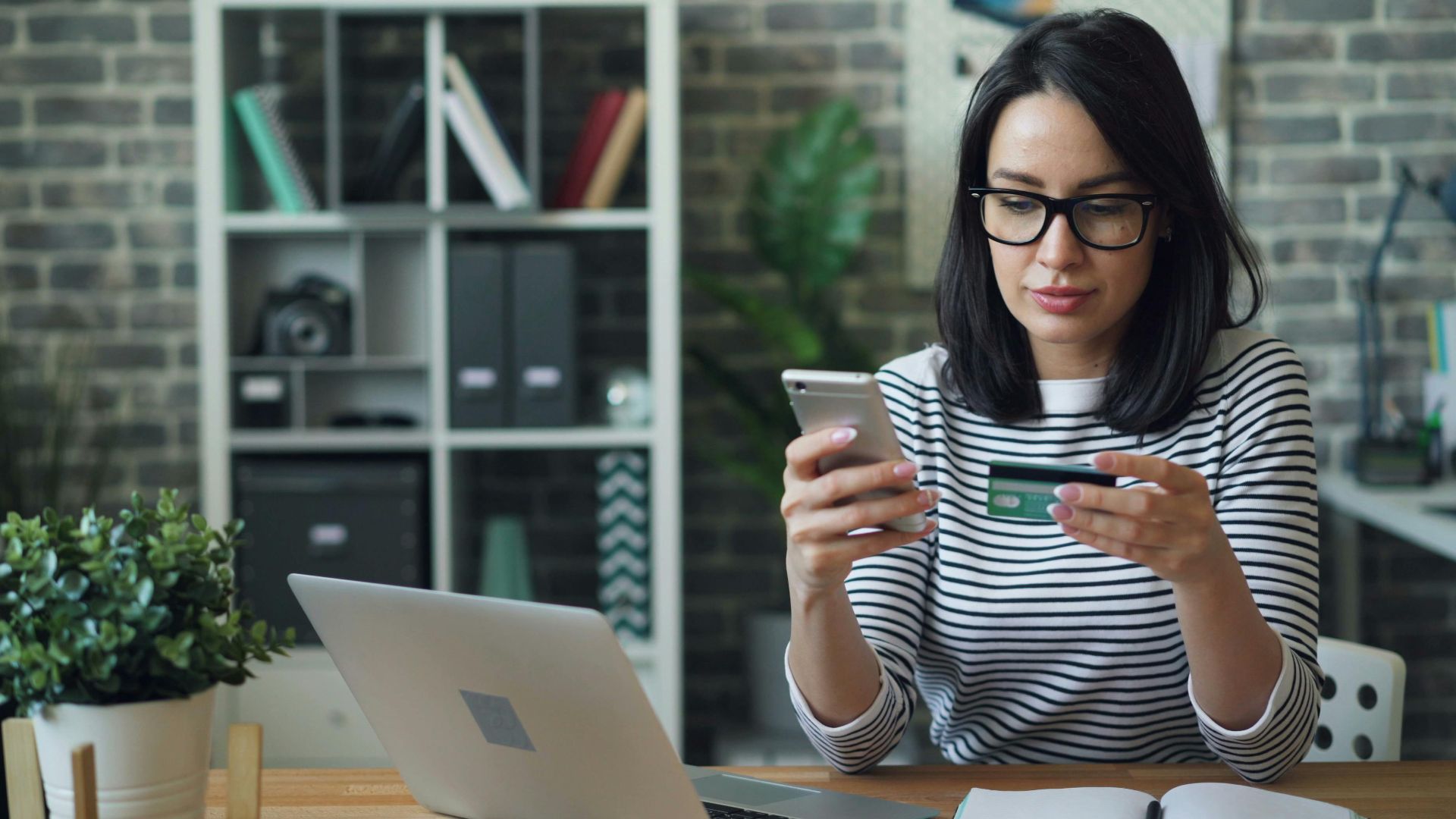 Professional woman using phone and credit card for online transaction at desk in office.
