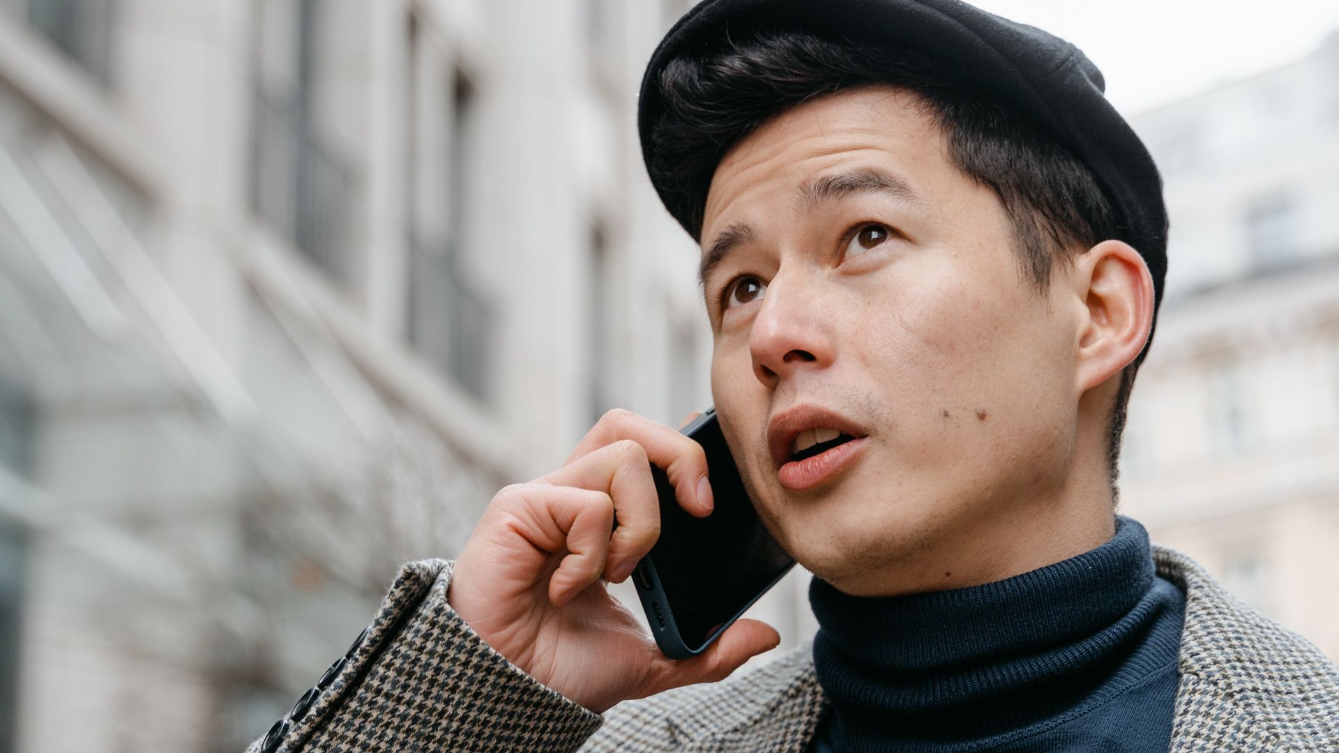 Stylish man in a city talking on a smartphone outdoors, wearing a coat and hat.