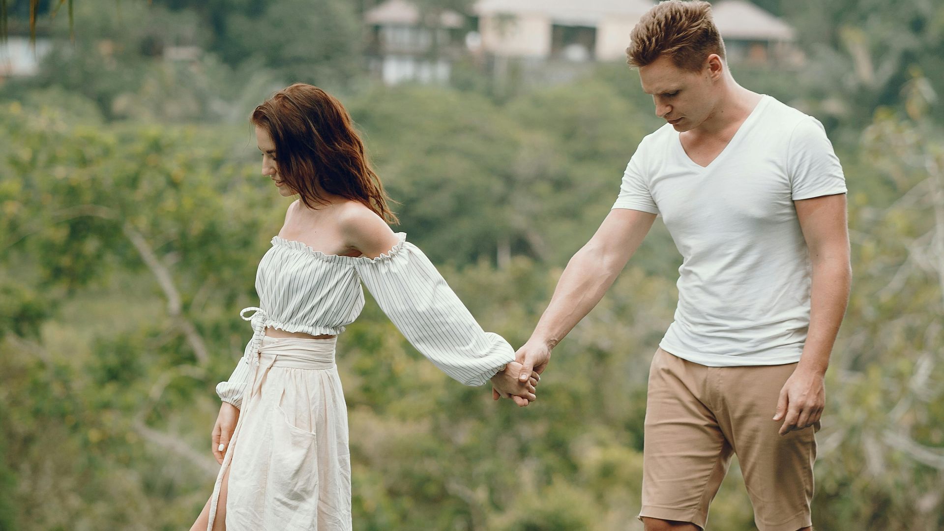 Full body barefoot anonymous stylish couple holding hands and standing on pool edge against scenic tropical countryside during romantic holidays