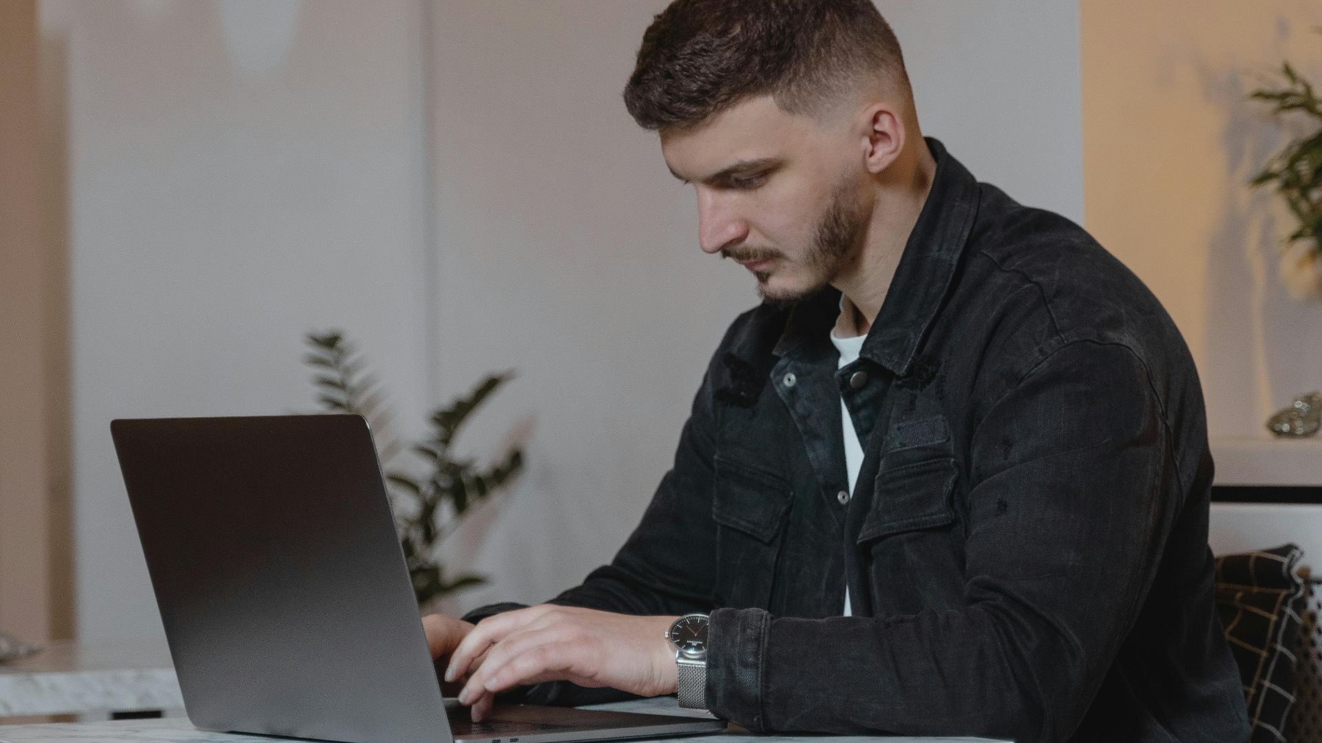 Young man in a denim jacket working on a laptop indoors, showcasing modern remote work lifestyle.