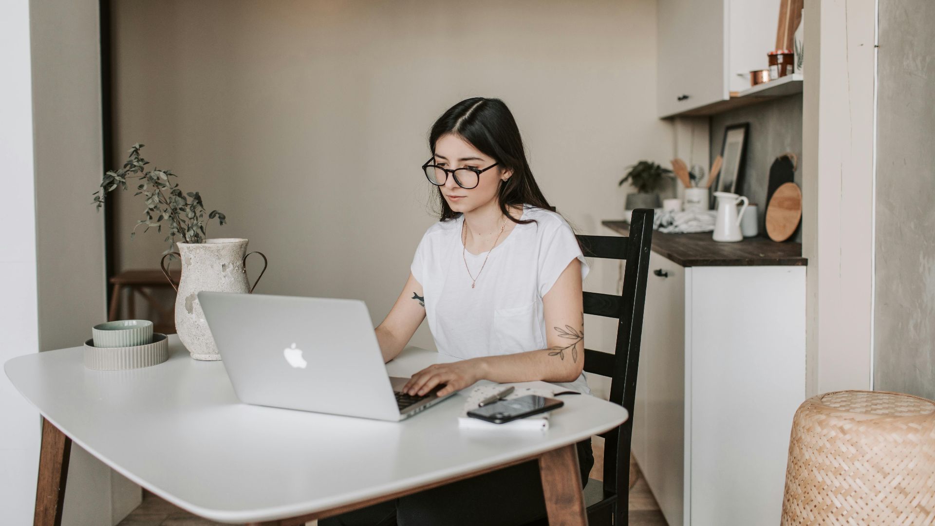 Smart female freelancer with tattooed arms wearing eyeglasses working on remote project browsing netbook while sitting at table with smartphone and notepad in modern room interior