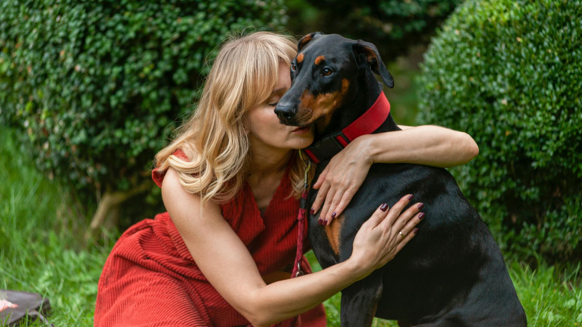 A woman in a red dress lovingly embraces her Doberman in a lush garden.