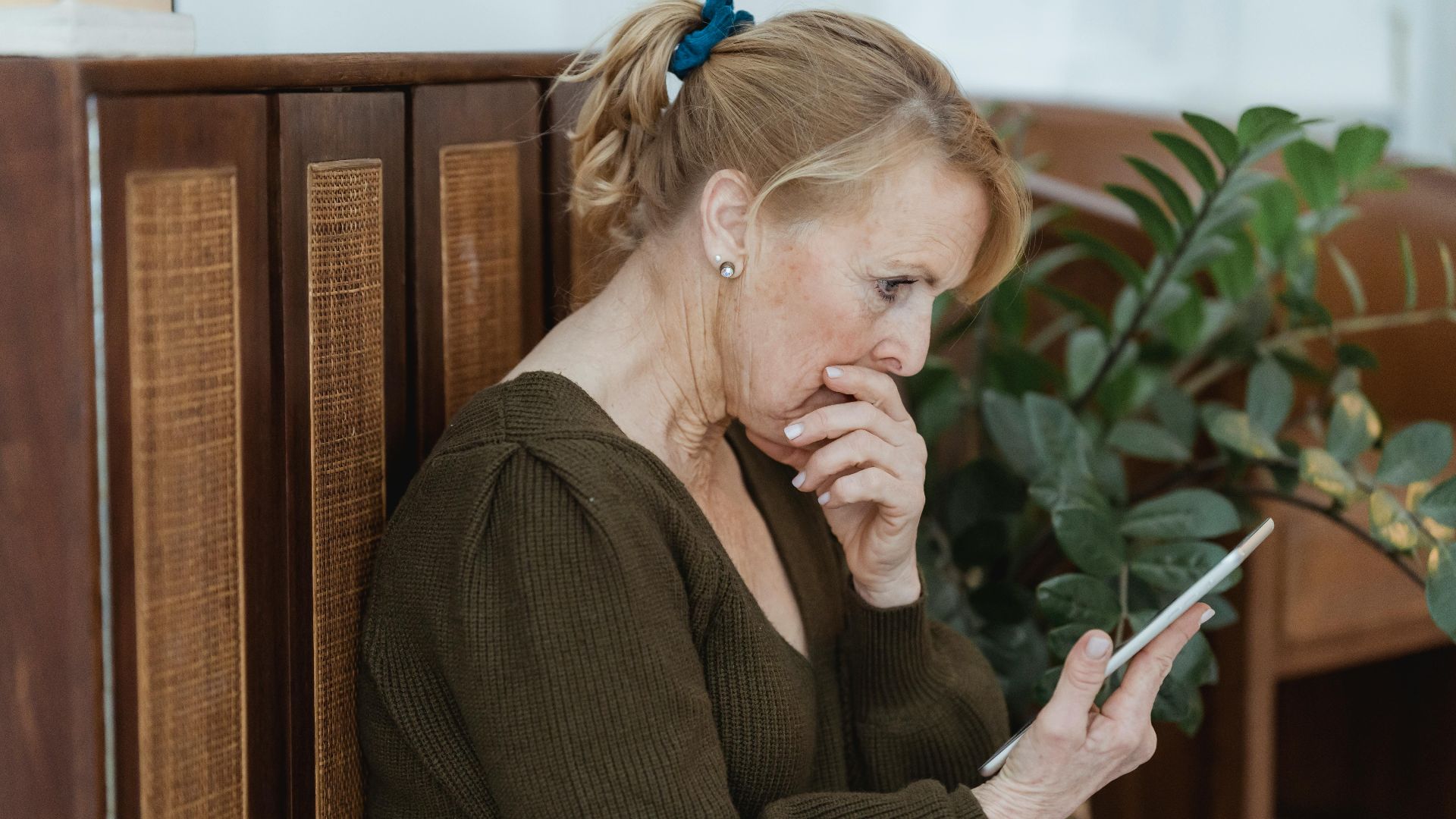 Side view of surprised elderly female with cellphone covering mouth against cabinet and plant at home