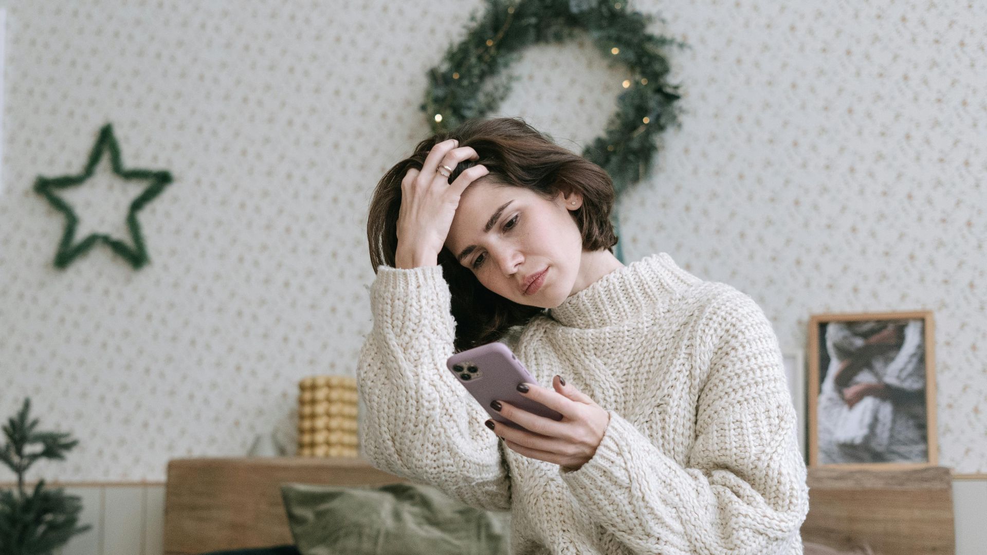 Woman in knitted sweater using smartphone while relaxing in a cozy bedroom.